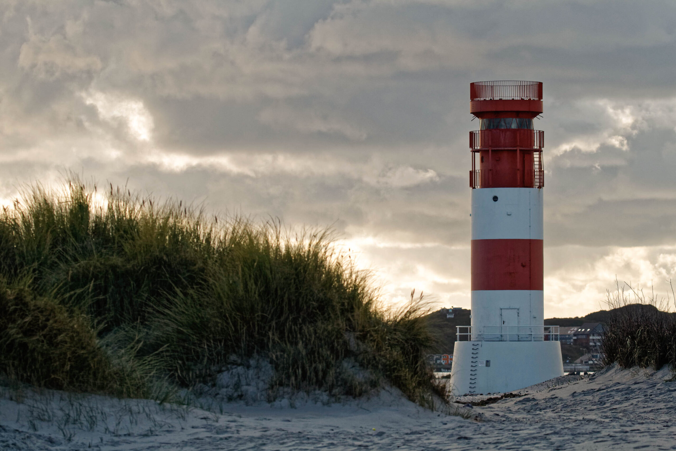 Das Bild zeigt einen Leuchtturm auf der Insel Helgoland. Der Leuchtturm ist weiß und rot gestreift und steht auf einem Sandhügel. Im Hintergrund sind Dünen und ein dramatischer Himmel mit Wolken zu sehen. Das Bild wurde im Oktober 2011 aufgenommen.