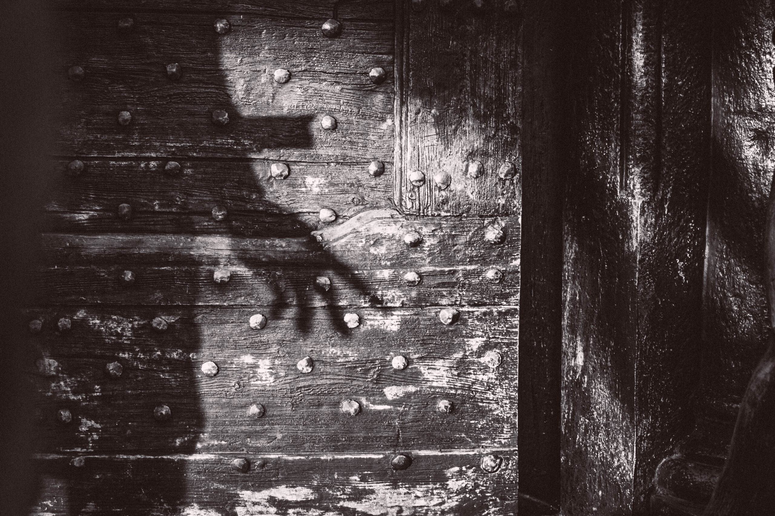 Das Foto zeigt eine detailreiche Nahaufnahme einer alten Holztür. Die Tür ist mit zahlreichen Nieten besetzt und weist deutliche Gebrauchsspuren und Verwitterung auf. Ein Schatten einer Hand ist auf der Türfläche sichtbar, was eine geheimnisvolle und fast unheimliche Atmosphäre erzeugt. Die Textur des Holzes und die Nieten sind sehr gut erkennbar, was die Detailtreue des Fotos unterstreicht. Die Beleuchtung ist dramatisch, mit starken Kontrasten zwischen Licht und Schatten, was die Stimmung des Bildes verstärkt. Der Hintergrund ist unscharf und trägt zur Fokussierung auf die Tür und den Schatten bei.