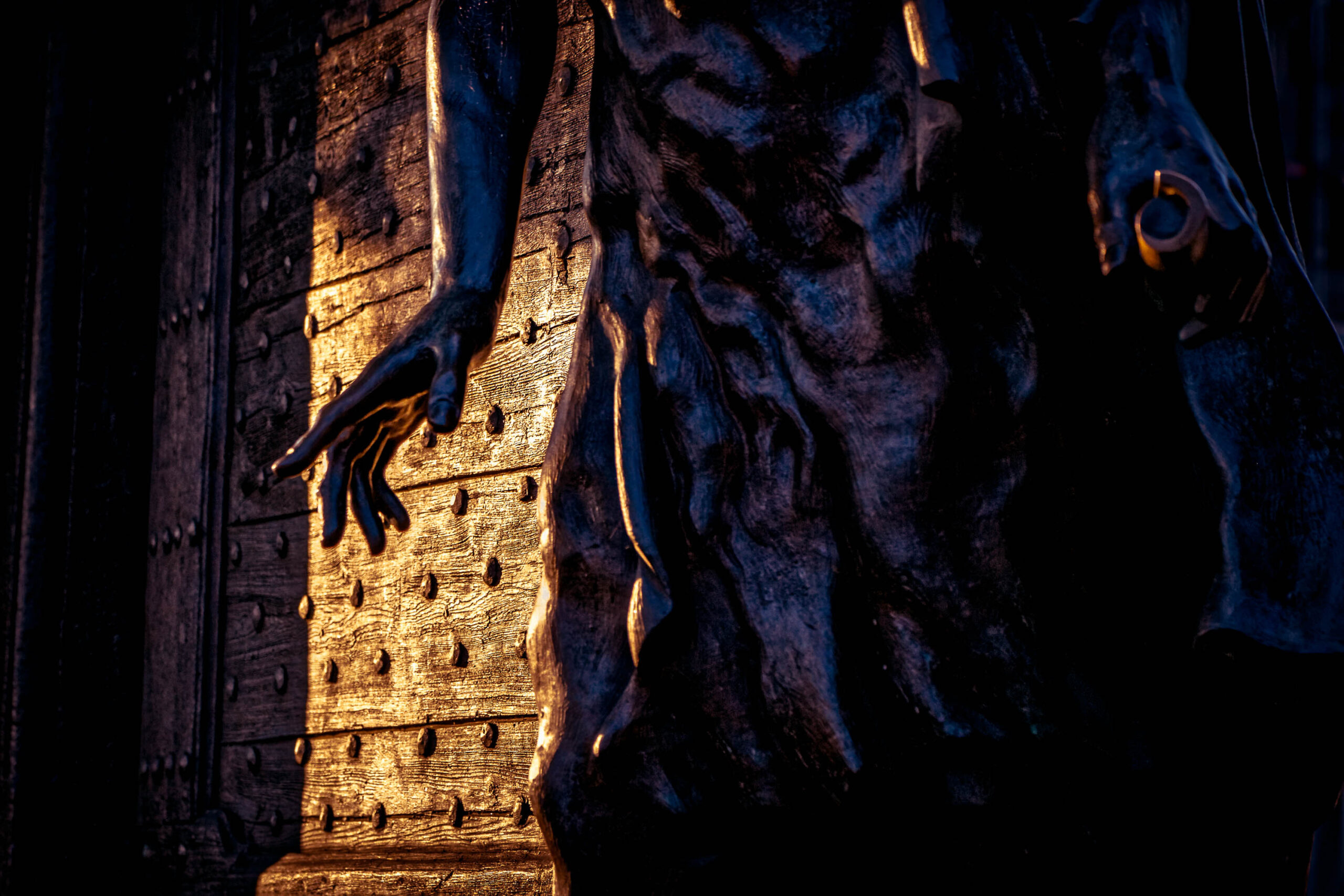 Die Aufnahme zeigt einen Detailbereich einer Bronzetür, vermutlich Teil eines größeren Kunstwerks oder einer architektonischen Struktur. Die Tür ist mit Reliefs versehen, die menschliche Hände, ein Gesicht und möglicherweise tierische Elemente darstellen. Das Licht fällt golden auf die Oberfläche, wodurch die Details der Skulpturen hervorgehoben werden und einen starken Kontrast zu den dunklen Bereichen der Tür bilden. Die Komposition ist auf die Textur und die künstlerische Gestaltung der Tür konzentriert, wobei der Hintergrund weitgehend unscharf ist, um die Aufmerksamkeit auf die Details zu lenken. Die Atmosphäre ist geheimnisvoll und künstlerisch, was durch die Beleuchtung und die Detailgenauigkeit der Skulpturen verstärkt wird.