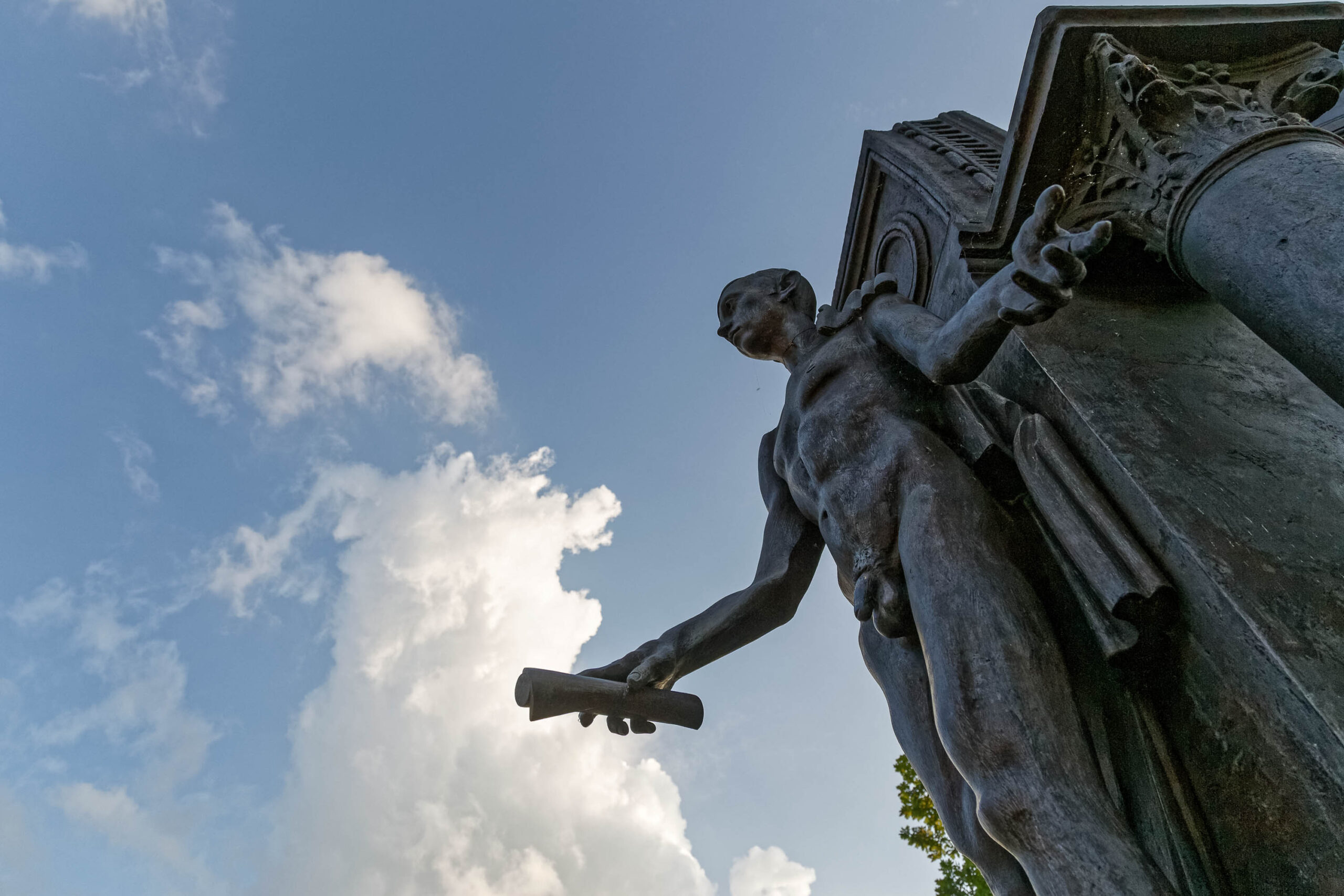 Das Foto zeigt einen Detailausschnitt einer monumentalen Bronzestatue, vermutlich Teil eines Denkmals oder einer Skulpturengruppe. Die Statue stellt eine männliche Figur in klassischer Robe dar, die ein Schriftrolle oder Dokument hält. Der Blickwinkel ist von unten aufgenommen, was die imposante Größe der Statue betont. Der Himmel im Hintergrund ist blau mit weißen Wolken, was eine sommerliche Atmosphäre schafft. Die Beleuchtung ist hell und betont die Details der Skulptur.