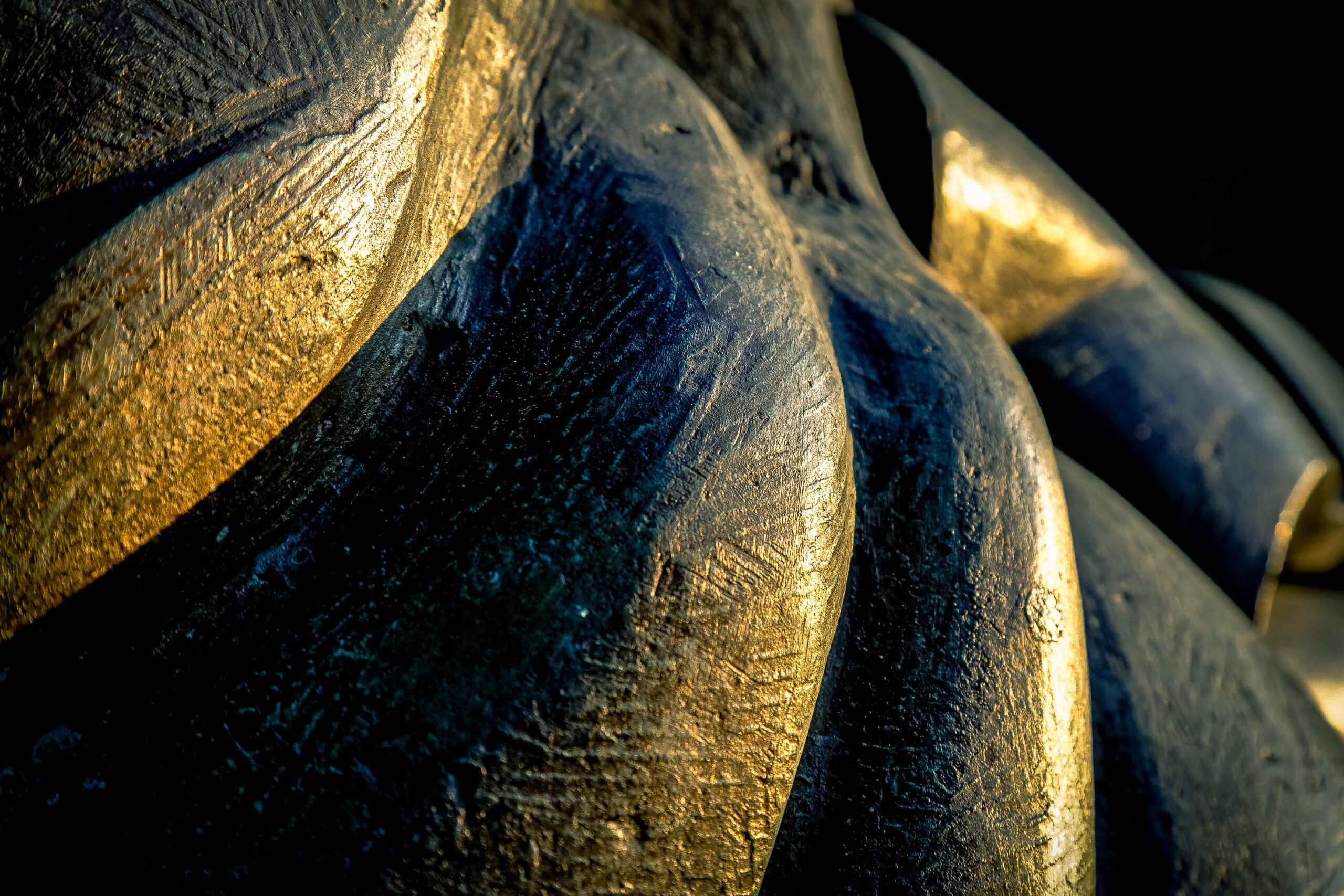 Die Aufnahme zeigt eine detaillierte Nahaufnahme einer Bronzestatue. Das Licht betont die Textur der Bronze und wirft dramatische Schatten, was eine fast abstrakte Wirkung erzeugt. Die Form scheint menschlich, ist aber durch die Detailgenauigkeit und den Fokus auf die Oberfläche schwer zu identifizieren. Die Statue ist in einem dunklen Umfeld platziert, was die goldenen Reflexionen der Bronze hervorhebt.