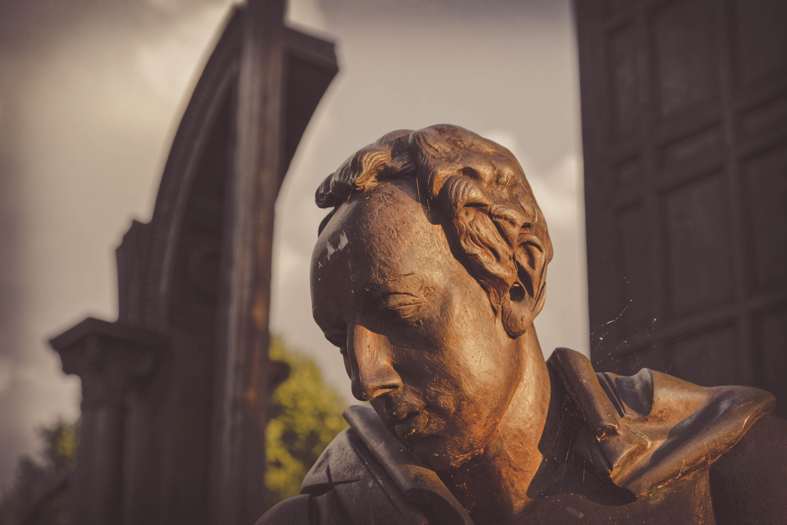 Die Fotografie zeigt eine detaillierte Nahaufnahme einer Bronzestatue eines Mannes. Das Gesicht des Mannes ist ernst und nachdenklich, mit einem leicht gesenkten Blick. Die Bronzefläche weist eine Patina auf, die dem Bild eine warme, erdige Tönung verleiht. Im Hintergrund ist ein Teil eines Gebäudes mit architektonischen Details und ein bewölkter Himmel erkennbar. Die Beleuchtung ist weich und diffus, was die Details der Statue hervorhebt und eine ruhige Atmosphäre schafft. Die Komposition konzentriert sich auf das Gesicht des Mannes, was den Betrachter dazu einlädt, über seine Gedanken und Gefühle nachzudenken.