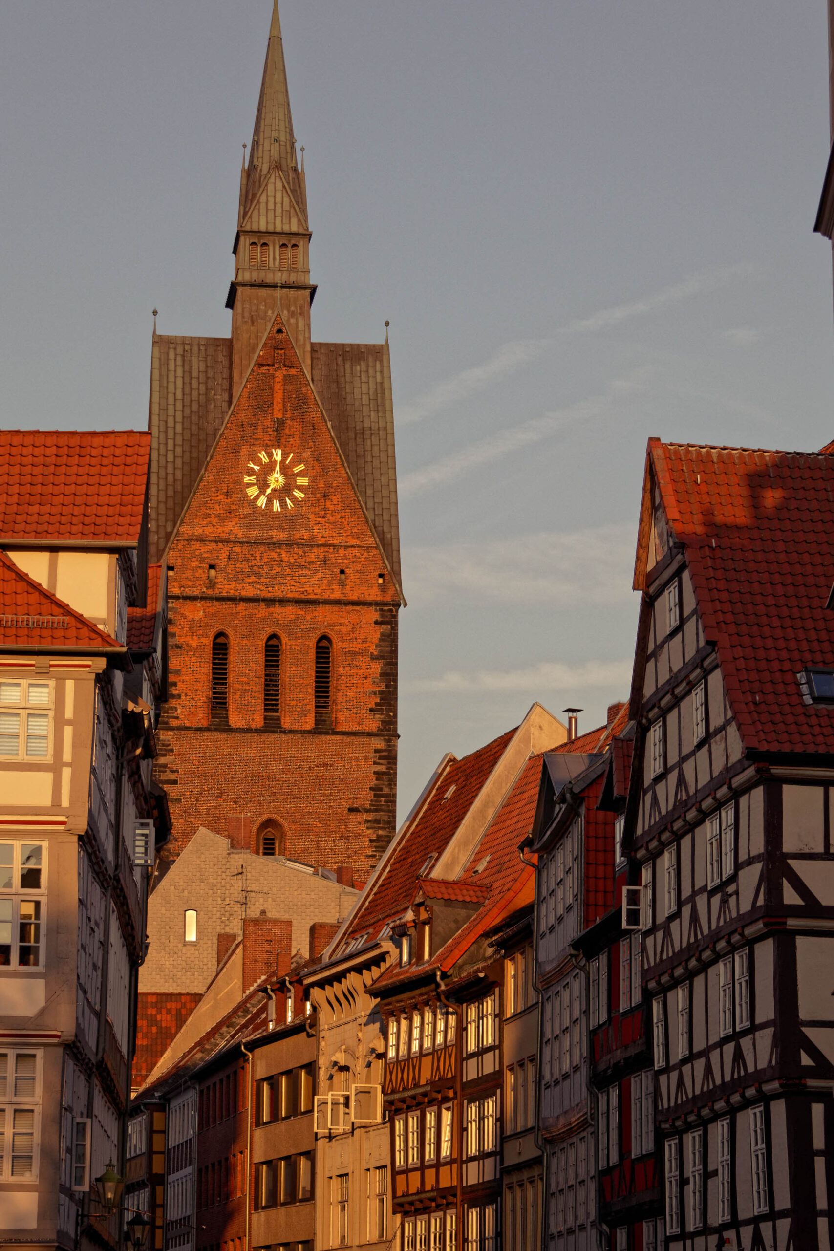 Das Foto zeigt eine malerische Ansicht der Altstadt von Hannover, Deutschland, bei Sonnenuntergang. Im Vordergrund stehen historische Fachwerkhäuser mit roten Ziegeldächern, die sich entlang einer schmalen Gasse ordnen. Im Hintergrund dominiert der Turm der Marktkirche, ein markantes Wahrzeichen der Stadt. Die warmen Farben des Sonnenuntergangs tauchen die Szene in ein goldenes Licht und verleihen ihr eine ruhige und besinnliche Atmosphäre.