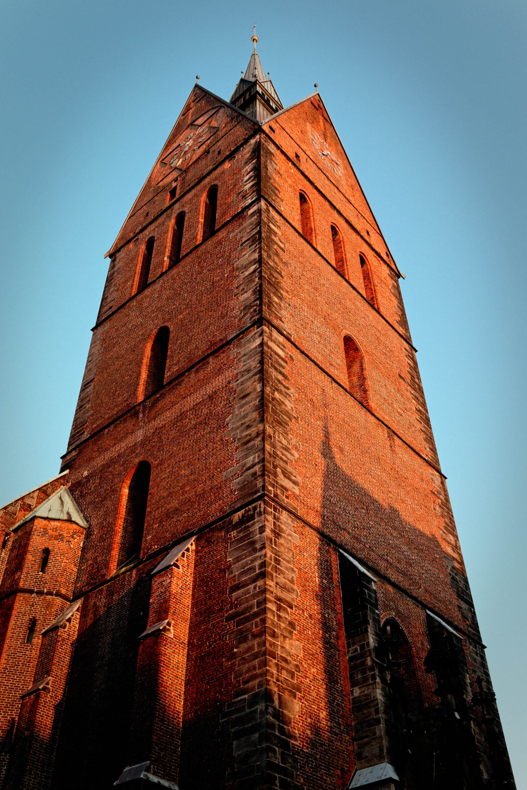 Das Foto zeigt eine Nahaufnahme des Turms der Marktkirche in Hannover, aufgenommen aus einem tiefen Blickwinkel, der die vertikale Ausrichtung und die Höhe des Turms betont. Der Turm besteht aus roten Backsteinen, die eine warme, erdige Farbe haben. Die Fassade ist mit gotischen Fenstern und dekorativen Elementen versehen. Der Himmel im Hintergrund ist blau und klar, was einen starken Kontrast zu den warmen Farben des Turms bildet. Das Licht scheint von der Seite zu kommen, was die Textur der Backsteine hervorhebt und Schatten wirft, die die Tiefe und Dimension des Turms betonen.