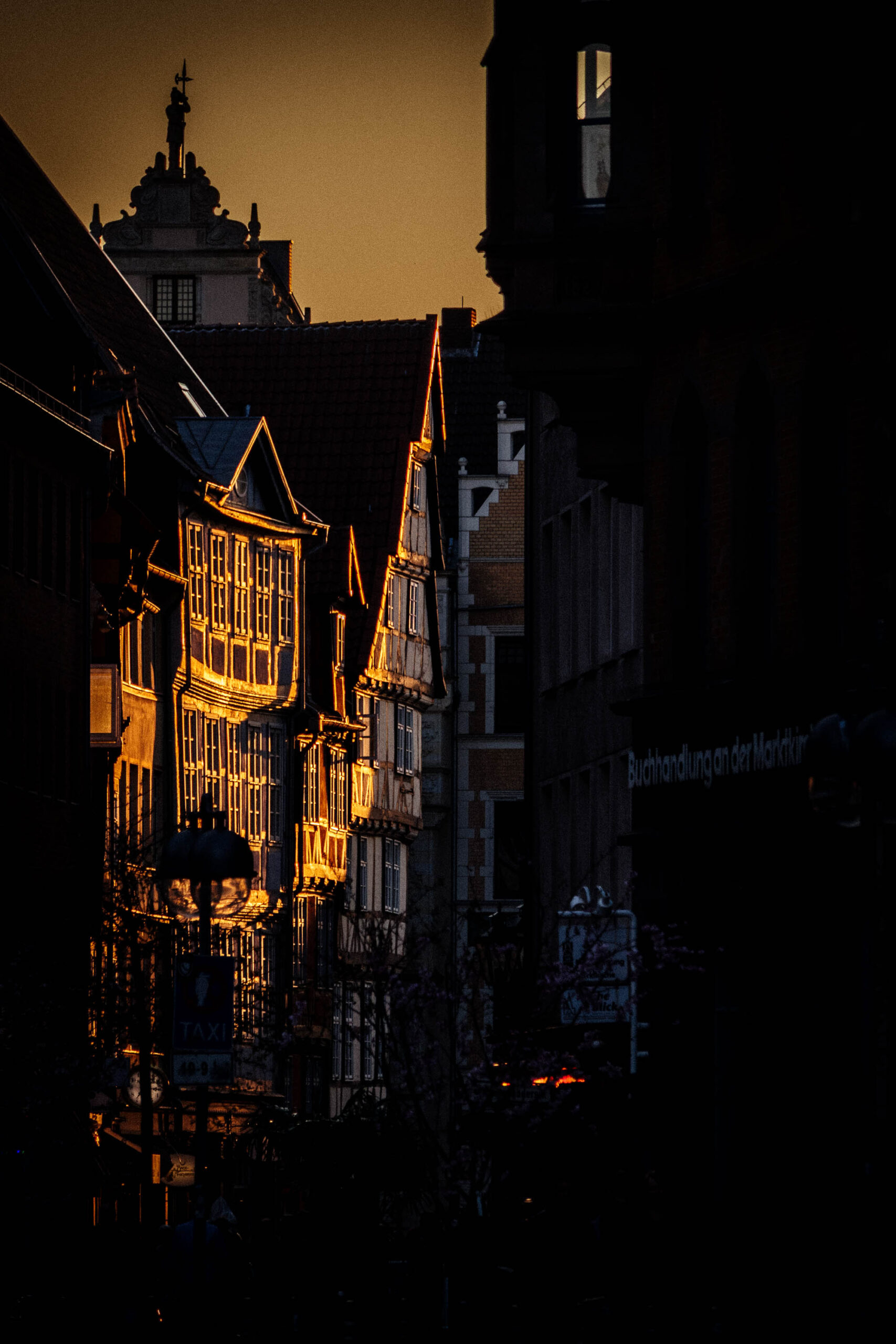 Die Aufnahme zeigt eine malerische Ansicht der Altstadt von Hannover bei Sonnenuntergang. Die Häuser, typisch für die historische Architektur der Stadt, sind in einem warmen, goldenen Licht getaucht, was eine stimmungsvolle und fast märchenhafte Atmosphäre erzeugt. Die Fassaden der Fachwerkhäuser sind detailliert erkennbar, wobei die Textur des Holzes und die architektonischen Details hervorgehoben werden. Im Vordergrund sind die Häuser mit ihren charakteristischen Dachformen und Fensteranordnungen zu sehen. Im Hintergrund ist ein höher gelegener Gebäuderiegel erkennbar, der die Perspektive der Aufnahme verstärkt. Der Himmel ist in warmen Farbtönen gehalten, was die romantische Stimmung der Aufnahme unterstreicht.