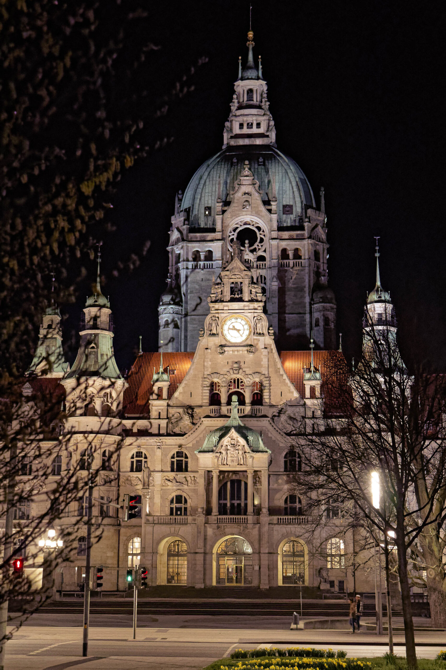 Das Foto zeigt das Neue Rathaus von Hannover bei Nacht. Die Beleuchtung betont die architektonischen Details des Gebäudes, insbesondere die Kuppel und die zahlreichen Türme und Erker. Im Vordergrund befindet sich eine gepflasterte Fläche mit einigen Blumenbeeten. Die Bäume im Vordergrund sind kahl und tragen zur stimmungsvollen Atmosphäre bei. Einige Personen sind im Bereich des Rathauses erkennbar.