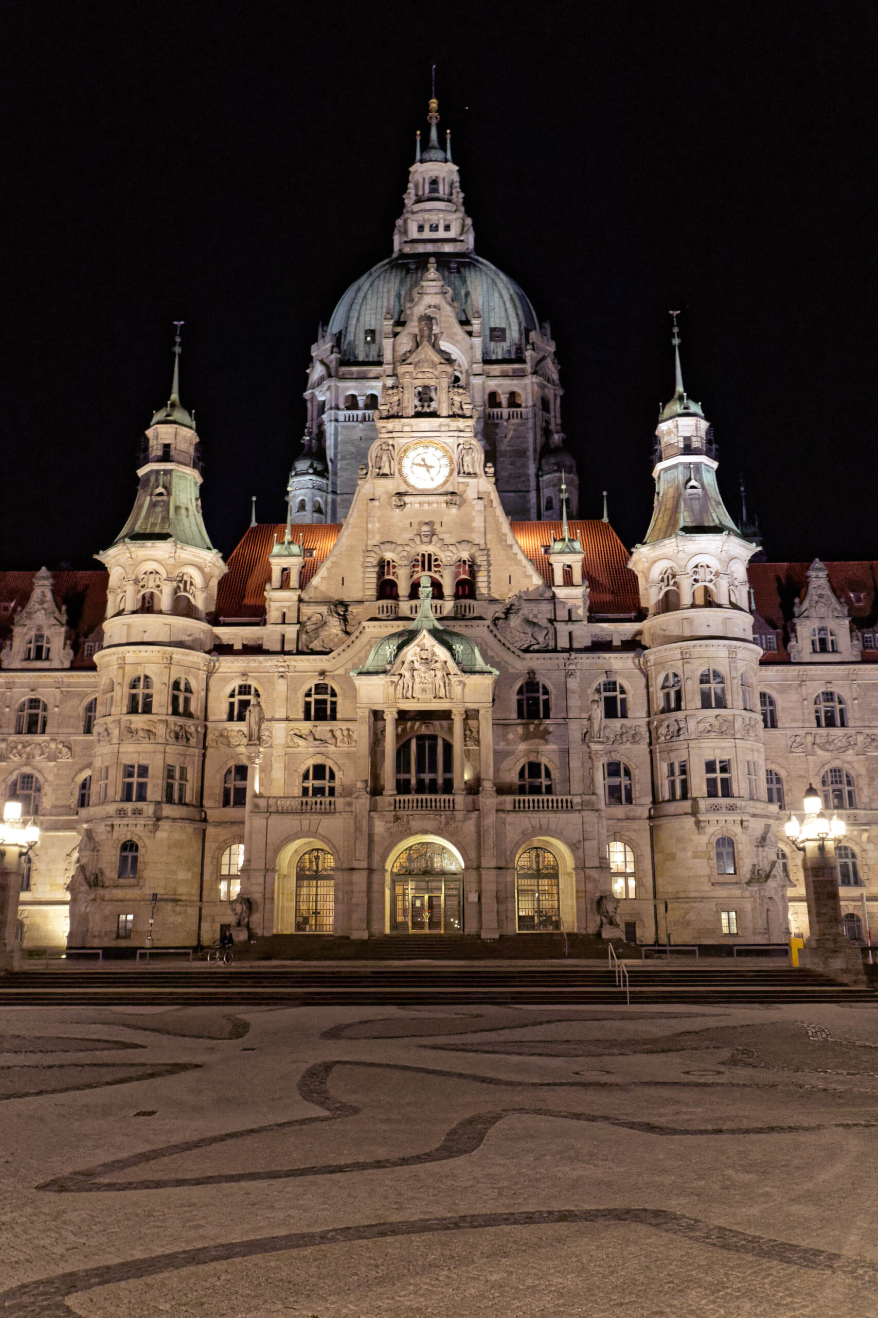 Das Foto zeigt das Neue Rathaus von Hannover bei Nacht. Das Rathaus ist im neorenaissance Stil erbaut und verfügt über eine beeindruckende Fassade mit zahlreichen Details, darunter Statuen, Erker und Türme. Die Beleuchtung betont die architektonischen Elemente und verleiht dem Gebäude eine majestätische Atmosphäre. Der Platz vor dem Rathaus ist leer und beleuchtet, was die Aufmerksamkeit auf das Gebäude lenkt. Der Himmel ist dunkel und klar, was die Beleuchtung des Rathauses hervorhebt.