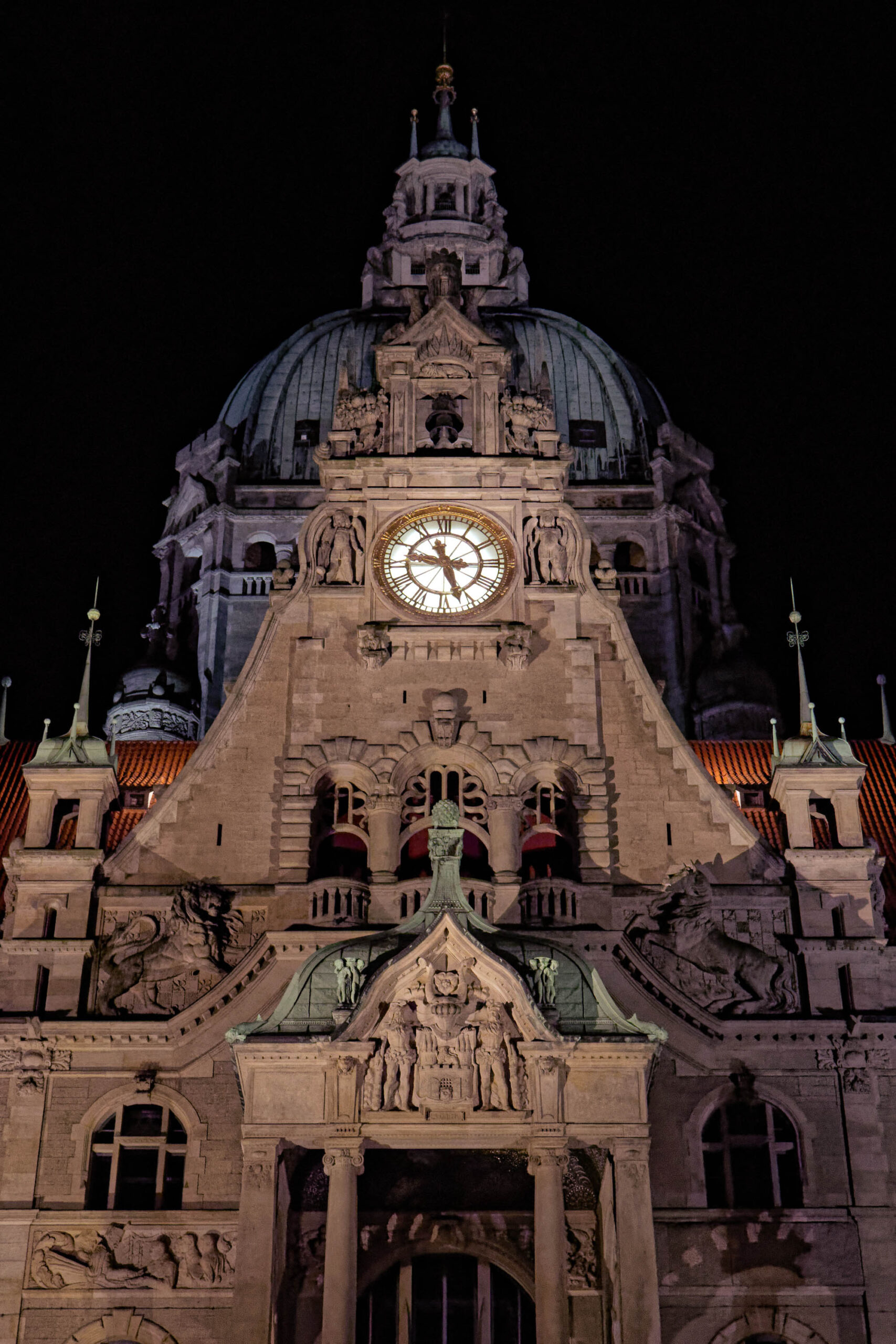 Das Foto zeigt eine detaillierte Ansicht des Neuen Rathauses in Hannover bei Nacht. Die Beleuchtung betont die architektonischen Details der Fassade, insbesondere die Skulpturen, Fenster und die Kuppel. Die Fassade ist im neugotischen Stil gehalten und zeigt eine Vielzahl von Ornamenten und Verzierungen. Die Kuppel ist mit einem kunstvollen Laternenaufbau gekrönt. Die Beleuchtung erzeugt eine dramatische und stimmungsvolle Atmosphäre.