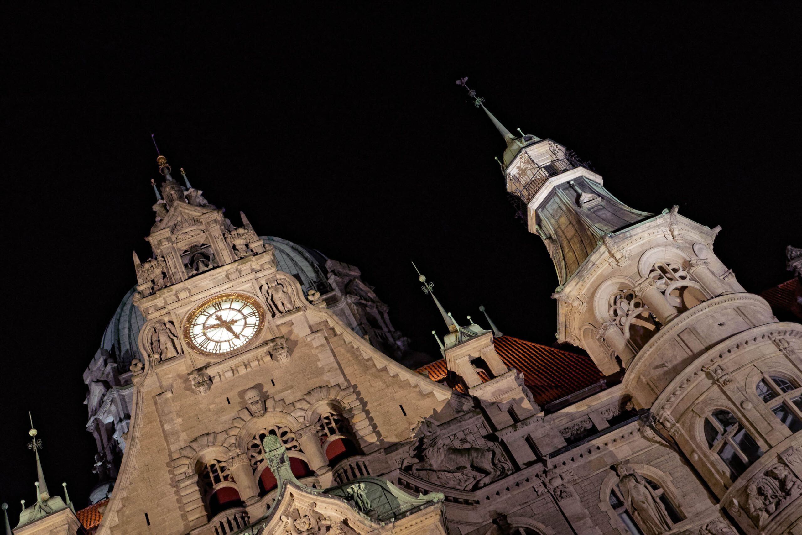 Das Foto zeigt eine nächtliche Ansicht der Marktkirche in Hannover, aufgenommen aus einem niedrigen Blickwinkel, der die beeindruckende Höhe und architektonische Details der Kirche hervorhebt. Die Beleuchtung betont die Fassade und die Türme, wobei die Uhr auf dem Hauptturm deutlich erkennbar ist. Der Himmel ist tiefschwarz, was die Kirche noch stärker hervorhebt.