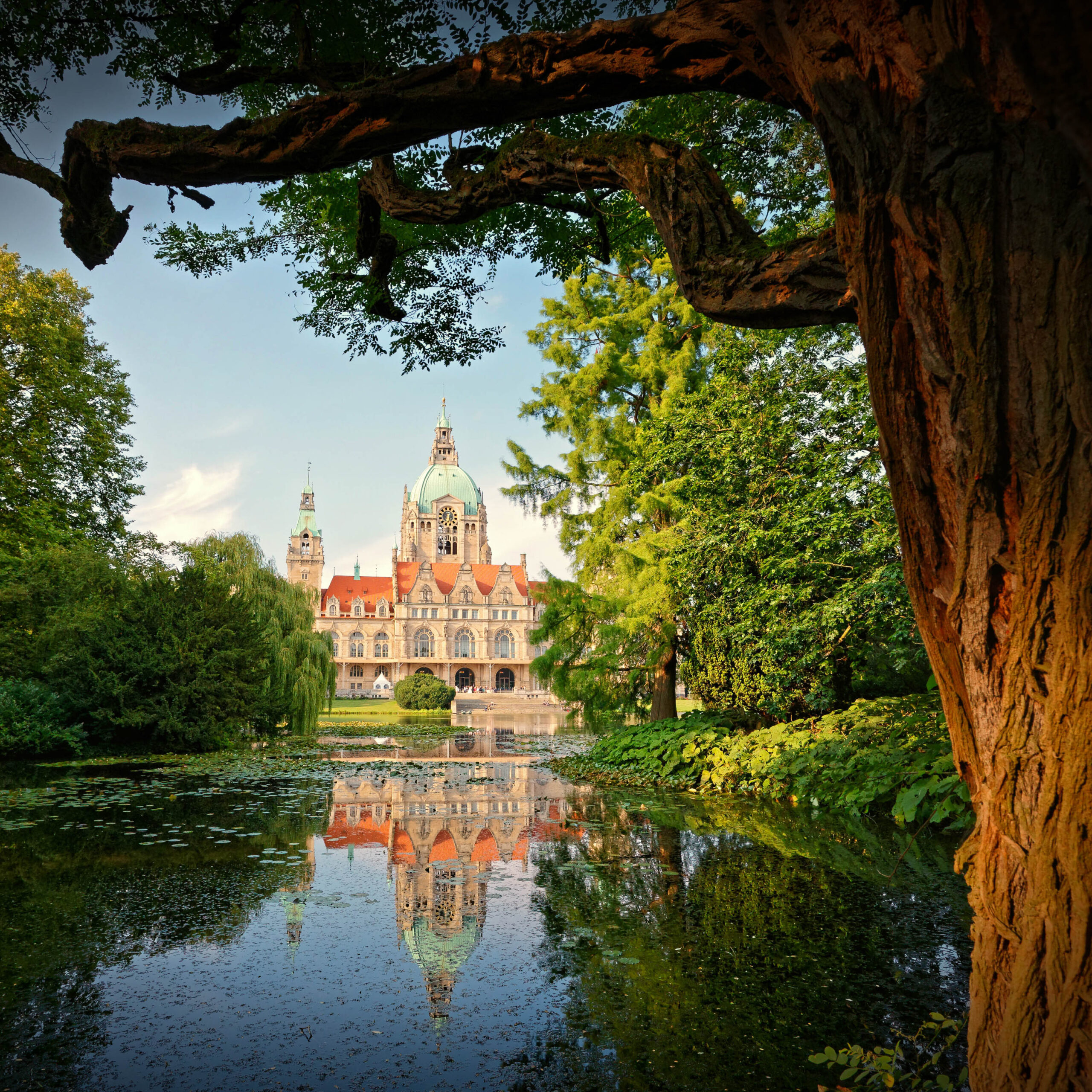 Das Foto zeigt eine malerische Ansicht des Herrenhäuser Schlosses in Hannover, eingebettet in die üppige Landschaft des Herrenhausengartens. Das Schloss, ein beeindruckendes Beispiel für den Stil des Barock, spiegelt sich in den ruhigen Gewässern eines Teichs wider, was eine symmetrische und fast surreale Komposition erzeugt. Die Spiegelung verstärkt die Pracht des Schlosses und verleiht dem Bild eine zusätzliche Dimension der Tiefe und Schönheit. Umgeben von dichtem Grün, das aus Bäumen und Sträuchern besteht, entsteht eine harmonische Verbindung zwischen Architektur und Natur. Das Licht ist weich und diffus, was die Farben des Schlosses und der Vegetation hervorhebt und eine friedliche Atmosphäre schafft.