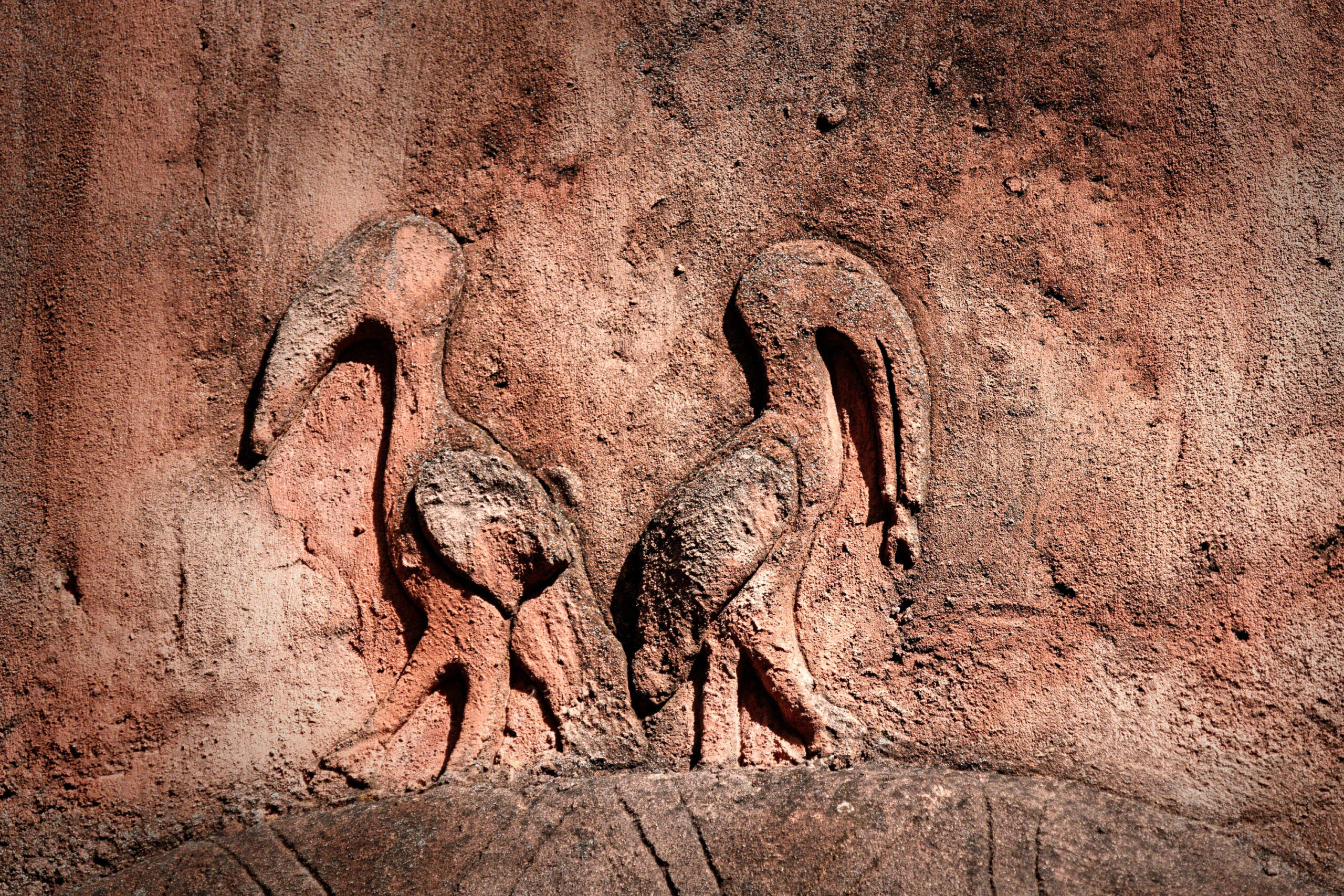 Das Bild zeigt eine Nahaufnahme von drei Elefanten-Skulpturen, die in eine Felswand-ähnliche Struktur eingelassen sind. Die Skulpturen stellen Elefanten in verschiedenen Posen dar: einer steht, einer beugt sich nach unten und der dritte scheint zu trinken oder zu fressen. Die Skulpturen sind in einem warmen, erdigen Farbton gehalten, der an eine natürliche Umgebung erinnert. Der Hintergrund ist rau und texturiert, was dem Bild eine organische und natürliche Atmosphäre verleiht. Die Beleuchtung ist weich und gleichmäßig, was die Details der Skulpturen hervorhebt und eine ruhige Stimmung erzeugt. Das Bild vermittelt ein Gefühl von Wildnis und Natur, obwohl es sich um eine künstliche Darstellung handelt. Es könnte Teil einer Diorama-Ausstellung oder einer thematischen Gestaltung in einem Zoo oder Museum sein.