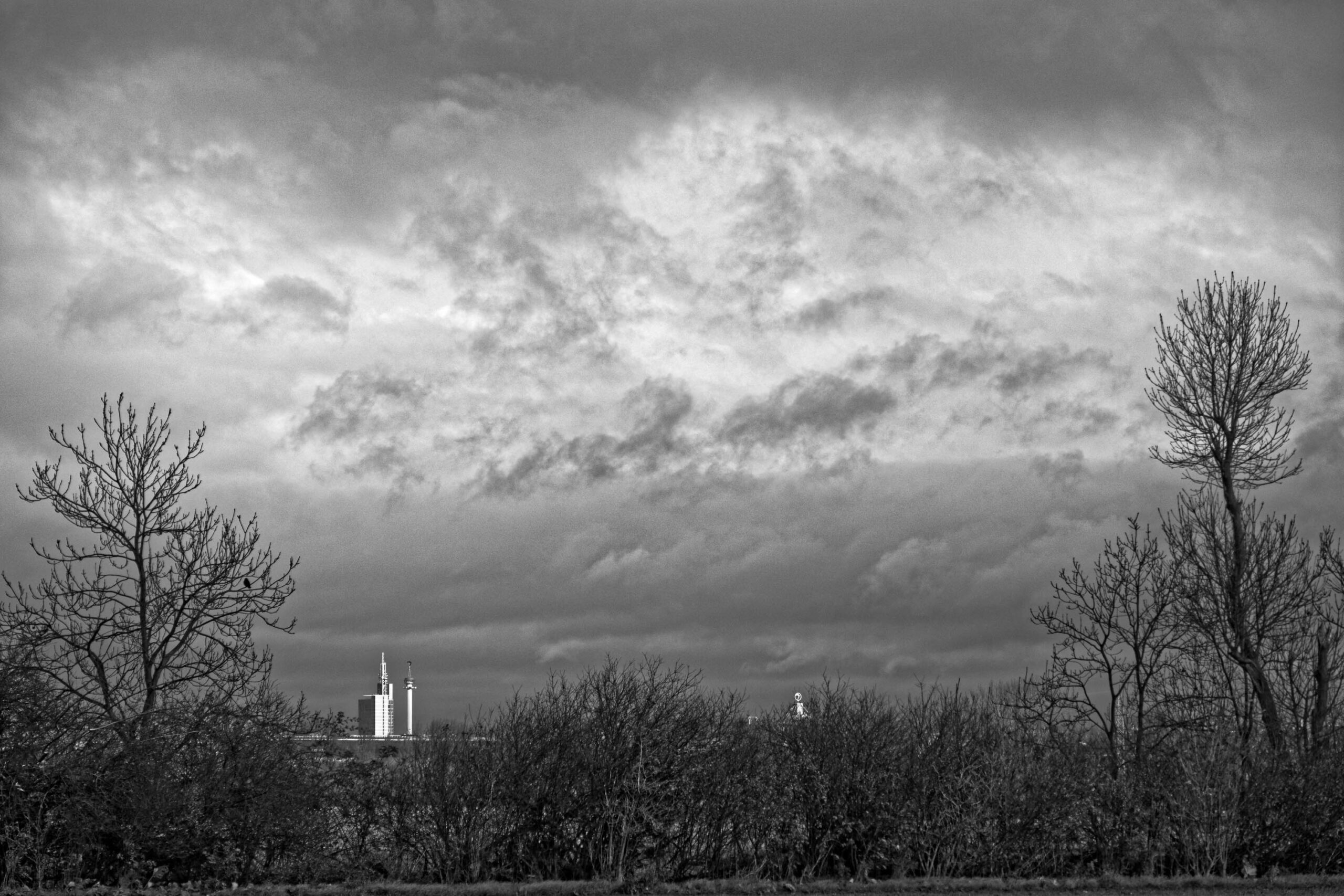 Das Foto zeigt eine düstere, monochrome Szene, die einen Blick auf ein markantes Gebäude im Hintergrund freigibt. Im Vordergrund dominieren kahle Bäume, die einen Rahmen um die Szene bilden. Der Himmel ist stark bewölkt, was die Szene zusätzlich düster wirken lässt. Die Perspektive ist von unten aufgenommen, was das Gebäude im Hintergrund hervorhebt. Die Landschaft wirkt verlassen und ruhig.