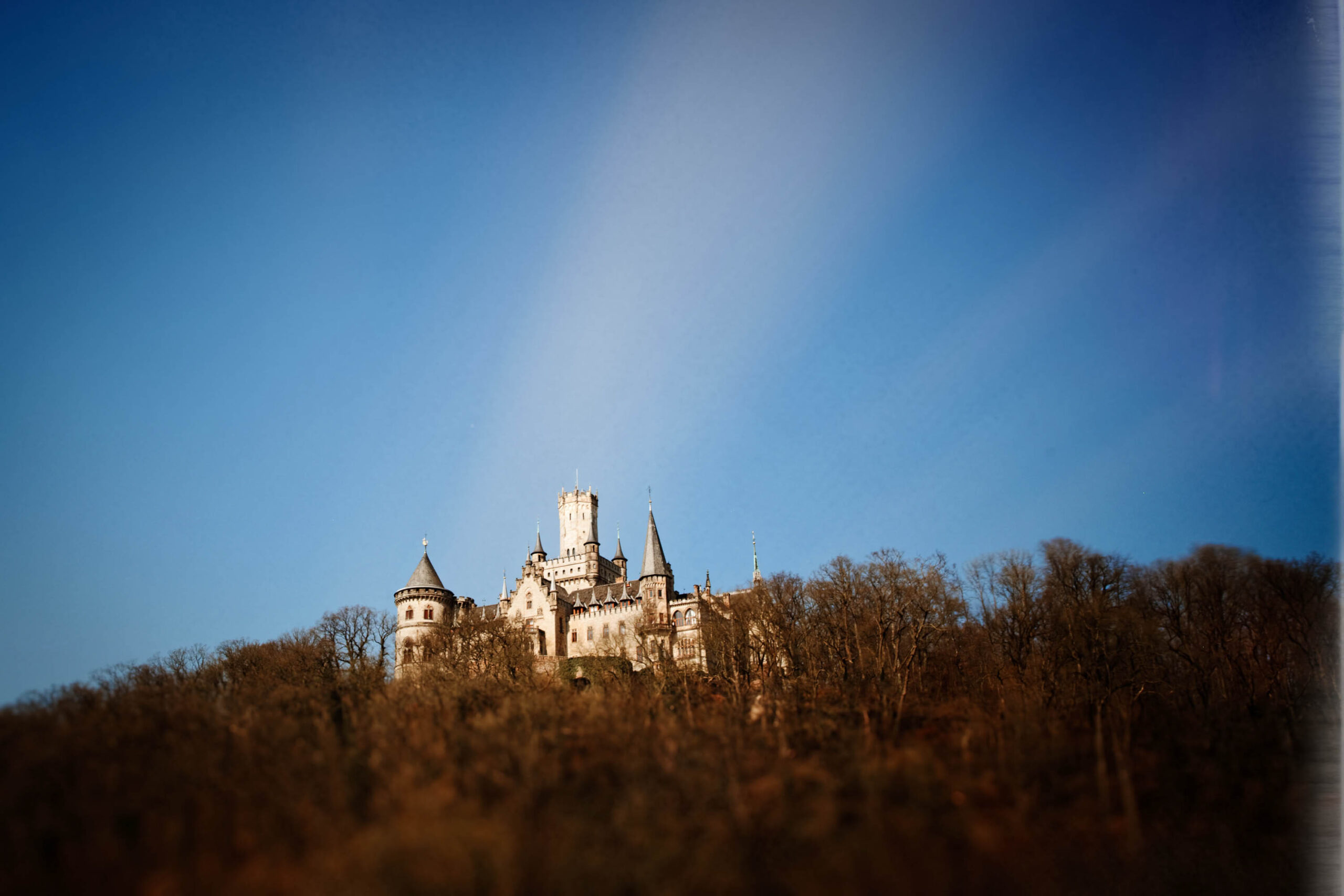 Das Foto zeigt das Schloss Pattensen, das sich auf einem Hügel inmitten eines dichten, kahlen Baumbestand thront. Der Himmel ist tiefblau und kontrastiert stark mit der hellen Fassade des Schlosses. Die Perspektive ist von unten aufgenommen, was die imposante Größe und die erhöhte Lage des Schlosses betont. Die Lichtverhältnisse sind gut, was die Details der Architektur hervorhebt.