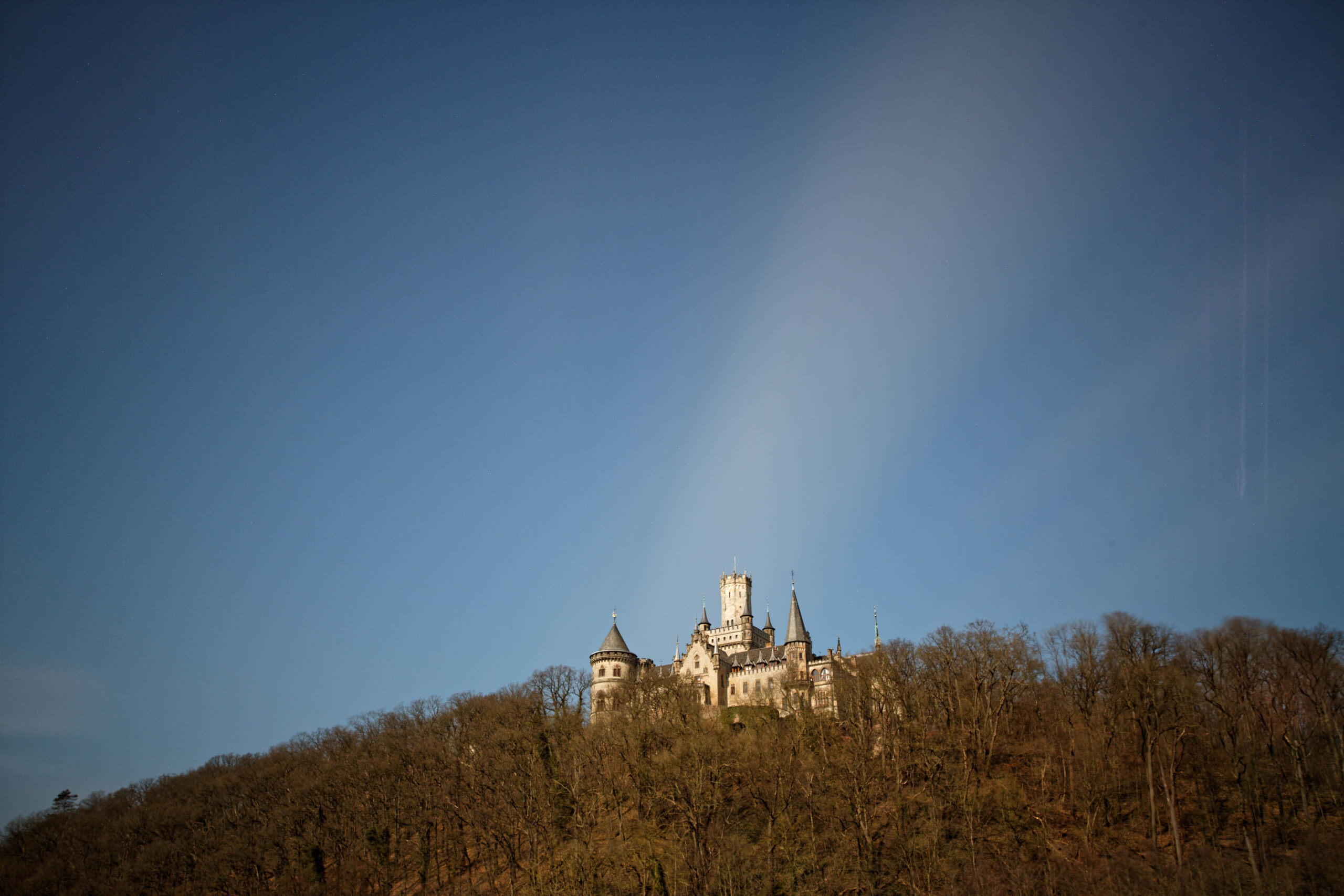 Das Foto zeigt das Schloss Kalenborn, das auf einem Hügel inmitten eines bewaldeten Geländes thront. Der Himmel ist tiefblau und klar, was die Farben des Schlosses und der umliegenden Landschaft hervorhebt. Das Schloss selbst ist im Detail gut erkennbar, mit seinen Türmen, Zinnen und dem markanten Erscheinungsbild. Die Bäume im Vordergrund sind kahl, was auf eine Winter- oder Übergangszeit hindeutet. Das Licht ist weich und gleichmäßig, was eine ruhige und friedliche Atmosphäre schafft.
