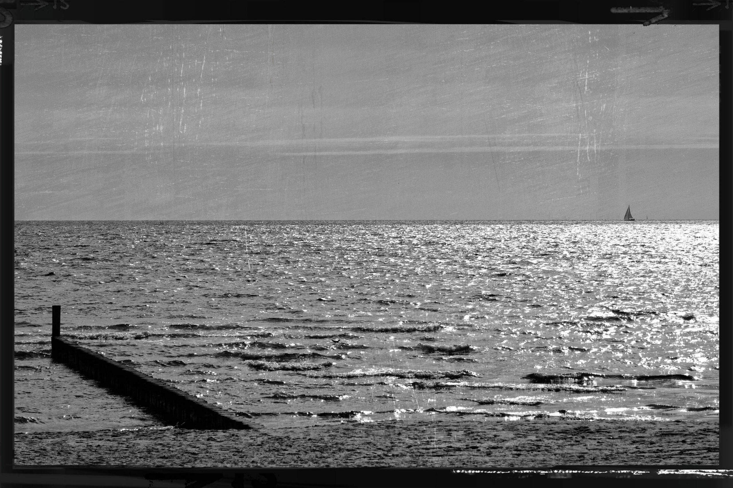Das Foto zeigt eine ruhige Szene an der Ostsee, aufgenommen in Ostholstein. Das Sonnenlicht reflektiert auf der Wasseroberfläche und erzeugt einen glitzernden Effekt. Ein hölzerner Wellenbrecher erstreckt sich im Vordergrund, während ein einzelnes Segelboot am Horizont zu sehen ist. Die Szene vermittelt eine friedliche und ruhige Atmosphäre.