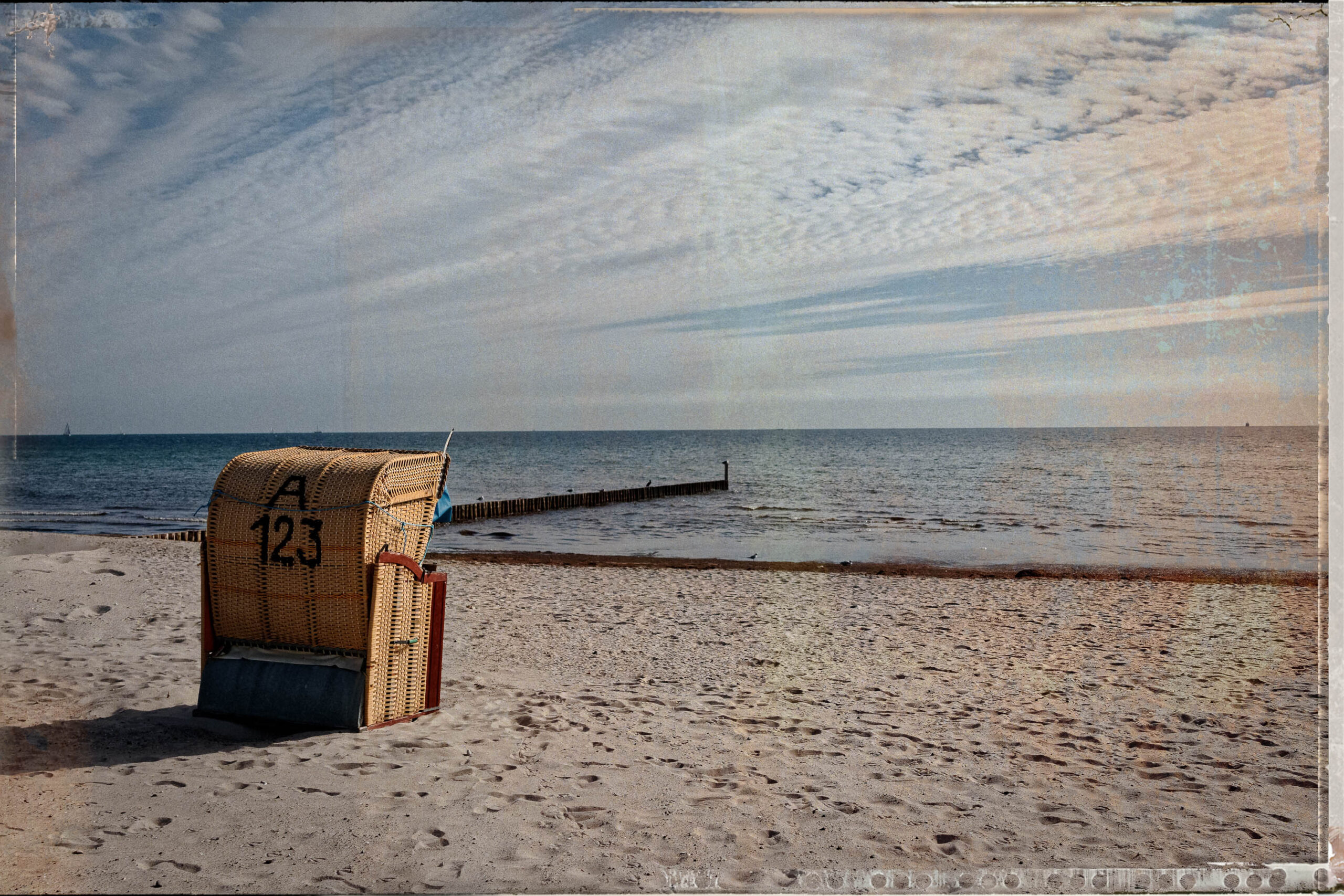 Das Bild zeigt einen typischen Strandkorb an einem Sandstrand an der Ostsee. Der Korb ist aus Weidengeflecht und hat die Nummer 'A 123' darauf. Im Hintergrund ist das Meer zu sehen, das in einem bläulichen Farbton erscheint. Ein hölzerner Steg ragt ins Wasser hinein. Der Himmel ist bewölkt, aber es scheint sonnig zu sein. Der Sand ist hell und reflektiert das Licht. Die Szene vermittelt eine ruhige und entspannte Atmosphäre.
