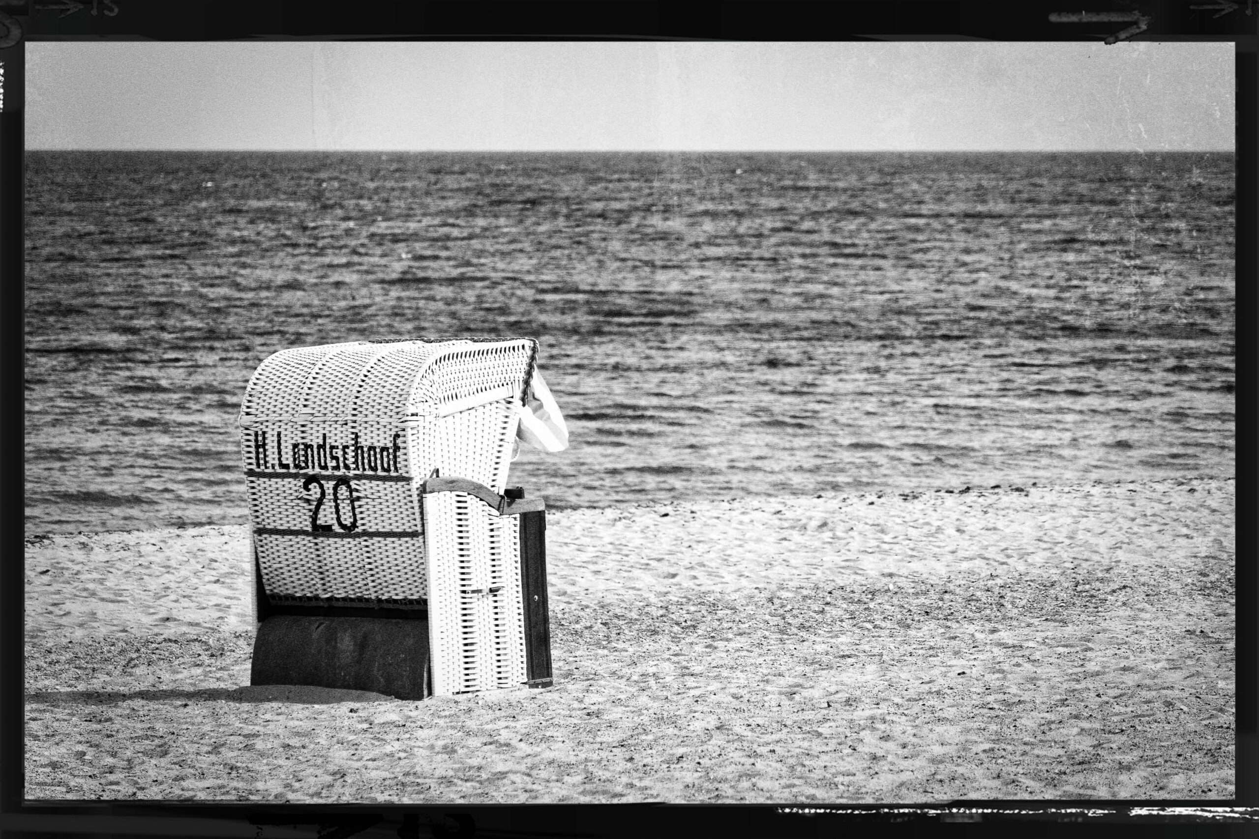 Das Foto zeigt einen traditionellen Korbsessel, der auf einem Sandstrand steht. Der Sessel ist aus geflochtenem Material gefertigt und weist eine typische Form auf, die in Küstenregionen Deutschlands anzutreffen ist. Im Hintergrund erstreckt sich das Meer, dessen Oberfläche durch leichte Wellenbewegung leicht strukturiert ist. Der Himmel ist nicht sichtbar, was den Fokus auf den Sessel und die Küstenlinie lenkt. Der Strand ist hell und sandig, was im Kontrast zum dunklen Sessel steht. Auf dem Sessel befindet sich ein Schild mit der Aufschrift 'Lüderslohe 20'. Das Bild vermittelt eine ruhige und entspannte Atmosphäre, typisch für einen Tag am Meer.