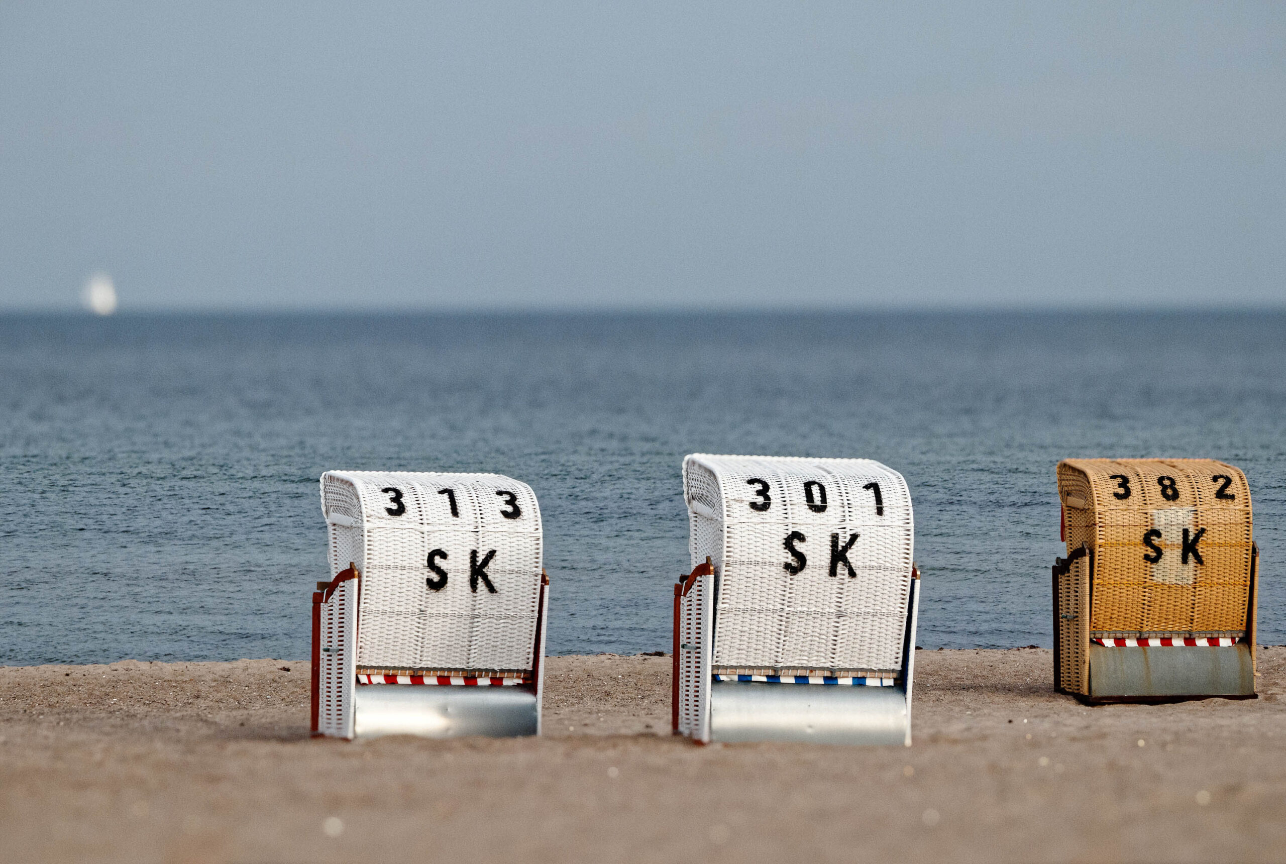 Das Bild zeigt drei traditionelle Strandkörbe, die auf einem Sandstrand stehen. Die Körbe sind aus Weidwerk gefertigt und mit roten und weißen Streifen verziert. Auf der Vorderseite jedes Korbs befindet sich ein Schild mit einer Nummer und dem Kürzel 'SK'. Im Hintergrund erstreckt sich das ruhige, blaue Meer, das bis zum Horizont reicht. Der Himmel ist leicht bewölkt und trägt zu einer ruhigen und entspannten Atmosphäre bei. Die Szene vermittelt ein Gefühl von Urlaub und Erholung an der Ostsee.
