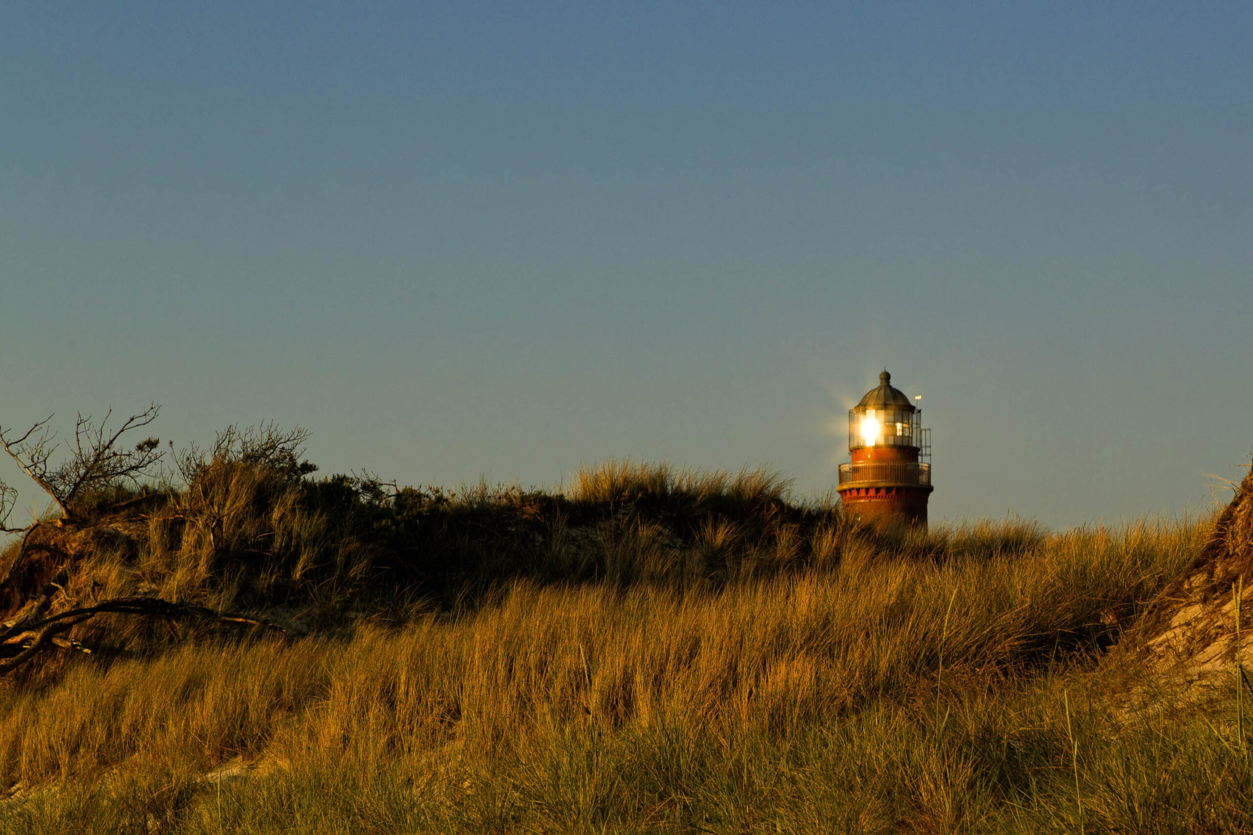 Die Aufnahme zeigt einen Leuchtturm, der sich inmitten einer Dünenlandschaft erhebt. Die Düne ist mit trockenem, golden-braunem Gras und vereinzelten Büschen bewachsen. Der Leuchtturm selbst ist rot gestrichen und besitzt eine gläserne Kuppel, die das Licht des Leuchtfeuers beherbergt. Der Himmel ist tiefblau und deutet auf den Abend oder die Morgendämmerung hin. Die Szene vermittelt eine ruhige und friedliche Atmosphäre.