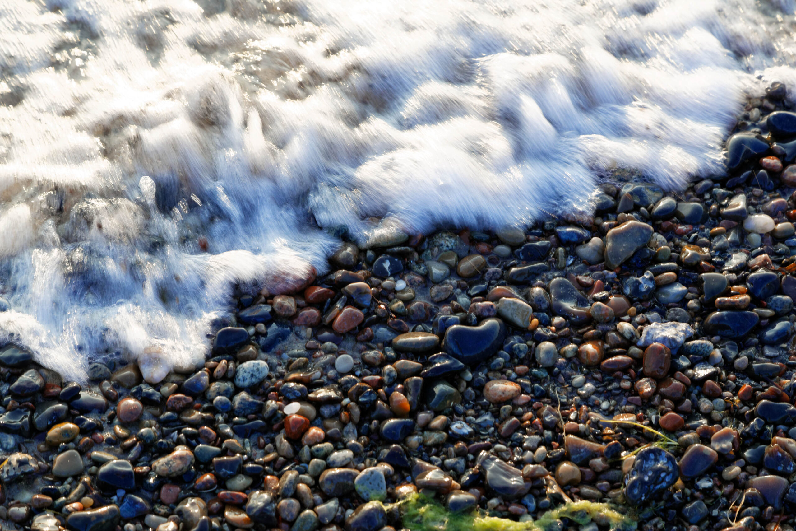 Die Aufnahme zeigt eine Detailaufnahme des Weststrandes auf der Halbinsel Darß. Das Bild konzentriert sich auf die Brandung, die auf einen Kieselstrand trifft. Die Wellen sind unscharf dargestellt, was die Bewegung des Wassers betont. Die Kieselsteine sind in verschiedenen Farben und Größen, was die Textur des Strandes hervorhebt. Das Licht ist hell und beleuchtet die Szene, was zu einem kontrastreichen Bild führt.