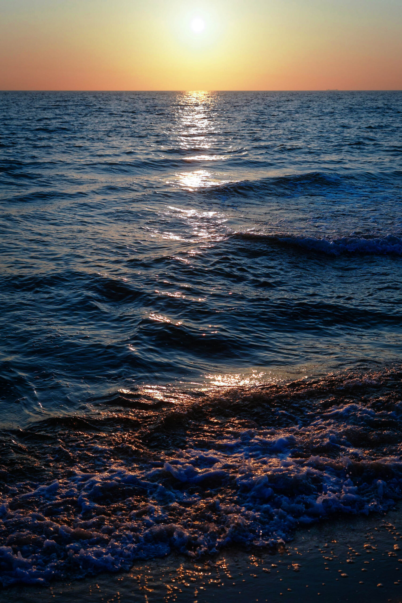 Das Bild zeigt einen Sonnenuntergang über der Ostsee, aufgenommen im Juni 2013 in Zingst. Das Wasser ist leicht aufgewühlt, mit kleinen Wellen, die an den Strand rollen. Ein heller Lichtpfad (Sonnenpfad) erstreckt sich über die Wasseroberfläche, der durch die untergehende Sonne entsteht. Der Himmel ist in warmen Farben von Orange und Gelb gehalten. Der Strand ist dunkel und sandig. Die Szene vermittelt eine ruhige und friedliche Atmosphäre.