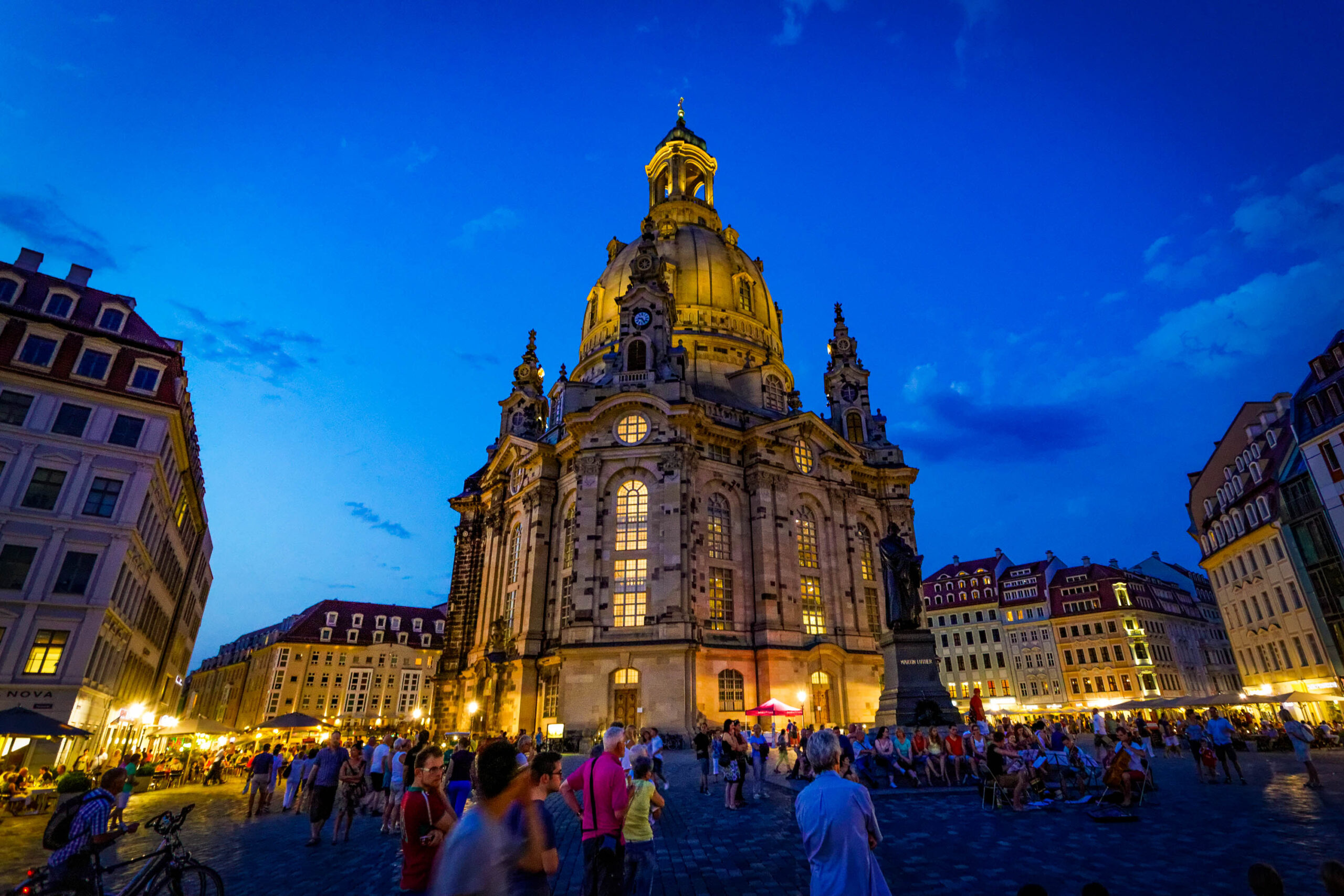Das Foto zeigt die Frauenkirche in Dresden bei Nacht. Die Kirche ist wunderschön beleuchtet, wobei die Kuppel und die Fassade in warmen Goldtönen erstrahlen. Um die Kirche versammelt sich eine große Menschenmenge, die die Atmosphäre genießen. Im Hintergrund sind weitere historische Gebäude zu sehen, die ebenfalls beleuchtet sind. Der Himmel ist dunkelblau und klar, was die Beleuchtung der Kirche und der Gebäude noch verstärkt. Das Foto fängt die Schönheit und den historischen Wert der Frauenkirche und ihrer Umgebung ein.