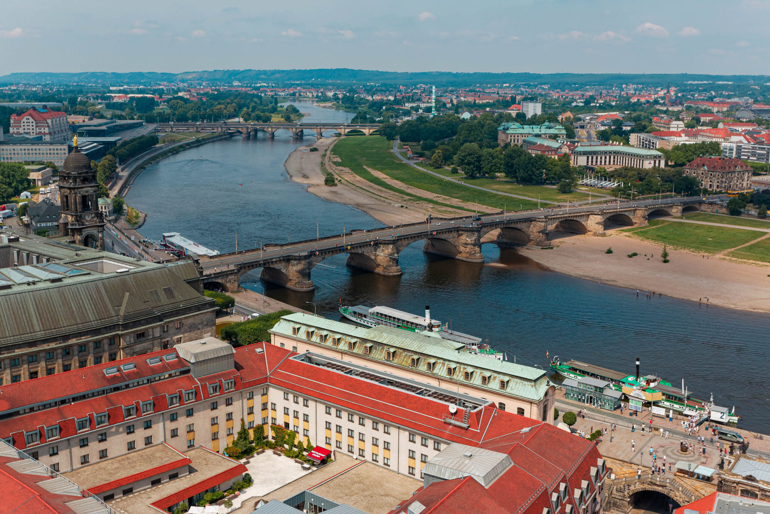 Die Aufnahme zeigt eine weitläufige Luftaufnahme von Dresden, aufgenommen im August 2013. Der Blick fällt auf die Elbe, die sich in einem sanften Bogen durch die Stadt schlängelt. Mehrere Brücken überspannen den Fluss, darunter die Augustusbrücke und die Waldschlößchenbrücke. Im Vordergrund sind die roten Ziegeldächer von Gebäuden in der Altstadt zu sehen. Im Hintergrund erstreckt sich das sächsische Elbtal mit seinen sanften Hügeln und Weinbergen. Der Himmel ist blau mit vereinzelten Wolken.