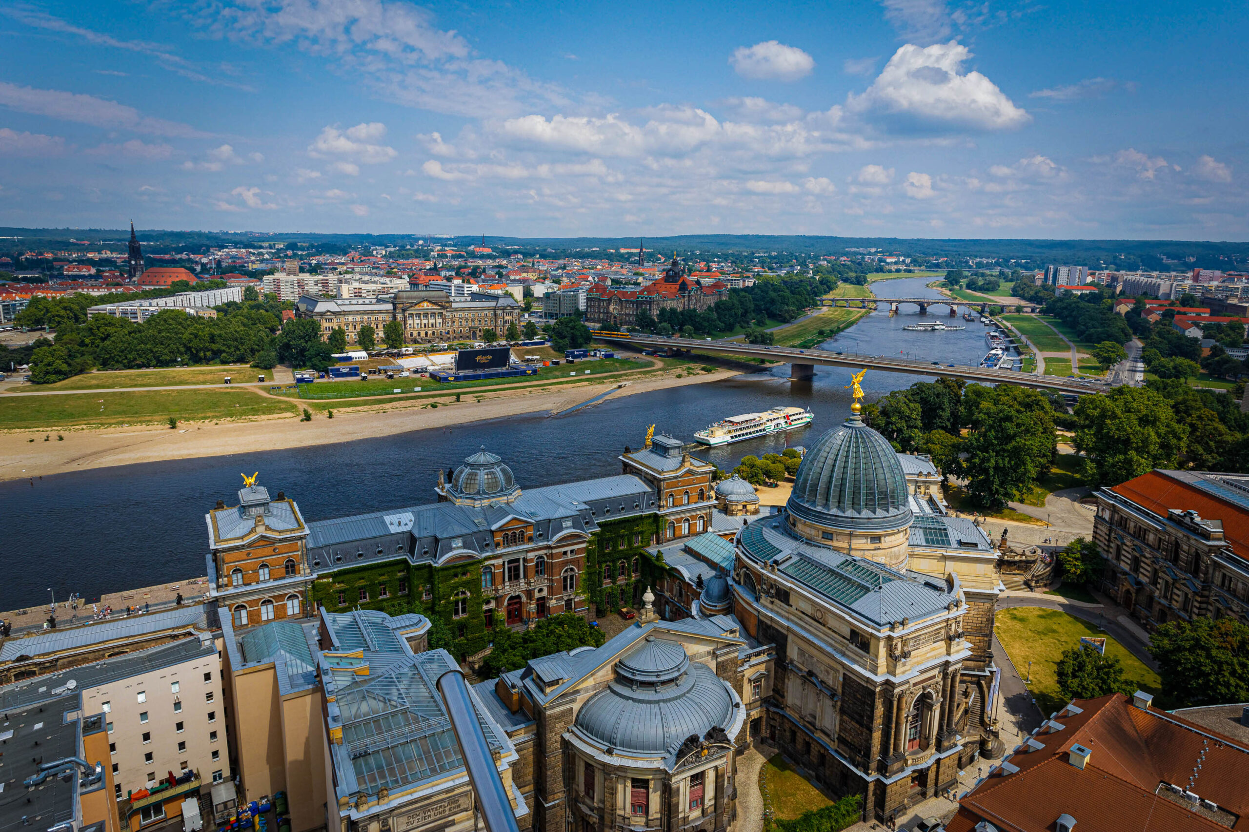 Die Aufnahme zeigt eine hochauflösende Vogelperspektive auf Dresden, Deutschland, aufgenommen im August 2013. Der Blick fällt auf den Zwinger-Palast, ein prächtiges Barockgebäude mit einer markanten Kuppel, der sich im Vordergrund befindet. Die Elbe schlängelt sich durch die Stadt und wird von mehreren Brücken überquert. Im Hintergrund erstreckt sich die Skyline von Dresden mit historischen Gebäuden und modernen Strukturen. Der Himmel ist blau mit vereinzelten weißen Wolken, was auf einen sonnigen Tag hindeutet. Die Perspektive bietet einen umfassenden Überblick über die Stadt und ihre Umgebung.