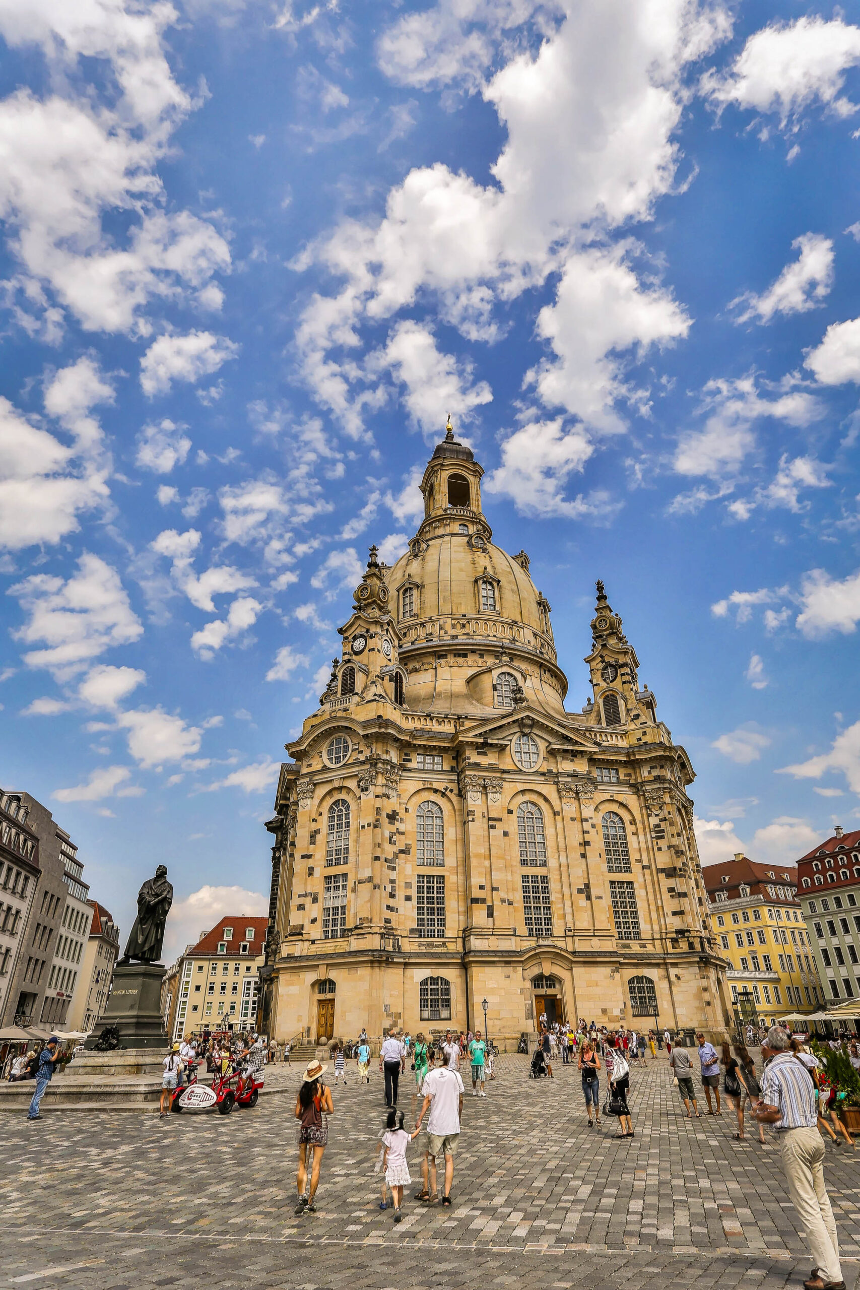 Das Foto zeigt die imposante Frauenkirche auf dem Neumarkt in Dresden. Die Kirche ist das zentrale Element des Bildes und dominiert die Szene mit ihrer goldenen Kuppel und den aufwändig verzierten Fassaden. Der Neumarkt ist mit Menschen gefüllt, die sich bewegen, entspannen und die Atmosphäre genießen. Der Himmel ist blau und von weißen Wolken durchzogen, was dem Bild eine helle und freundliche Stimmung verleiht. Im Hintergrund sind weitere Gebäude und Elemente der Stadt zu sehen, die den Kontext des Bildes vervollständigen. Das Foto wurde im August 2013 aufgenommen und fängt die Schönheit und den Wiederaufbau der Stadt Dresden ein.