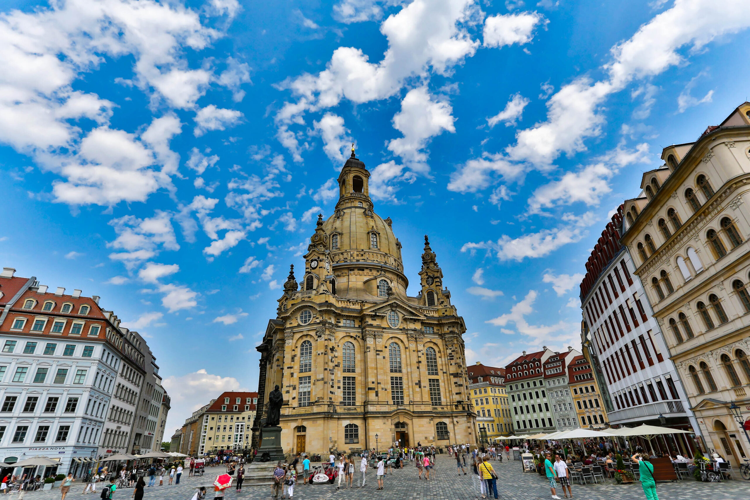 Das Bild zeigt die Frauenkirche in Dresden, Deutschland, aufgenommen im August 2013. Die Kirche dominiert die Szene und wird von umliegenden historischen Gebäuden flankiert. Der Neumarkt ist mit Menschen gefüllt, die sich entspannen, essen oder die Sehenswürdigkeit bewundern. Der Himmel ist blau mit weißen Wolken, was eine angenehme Atmosphäre schafft. Die Architektur der Kirche und der umliegenden Gebäude ist beeindruckend und zeugt von der Geschichte der Stadt.