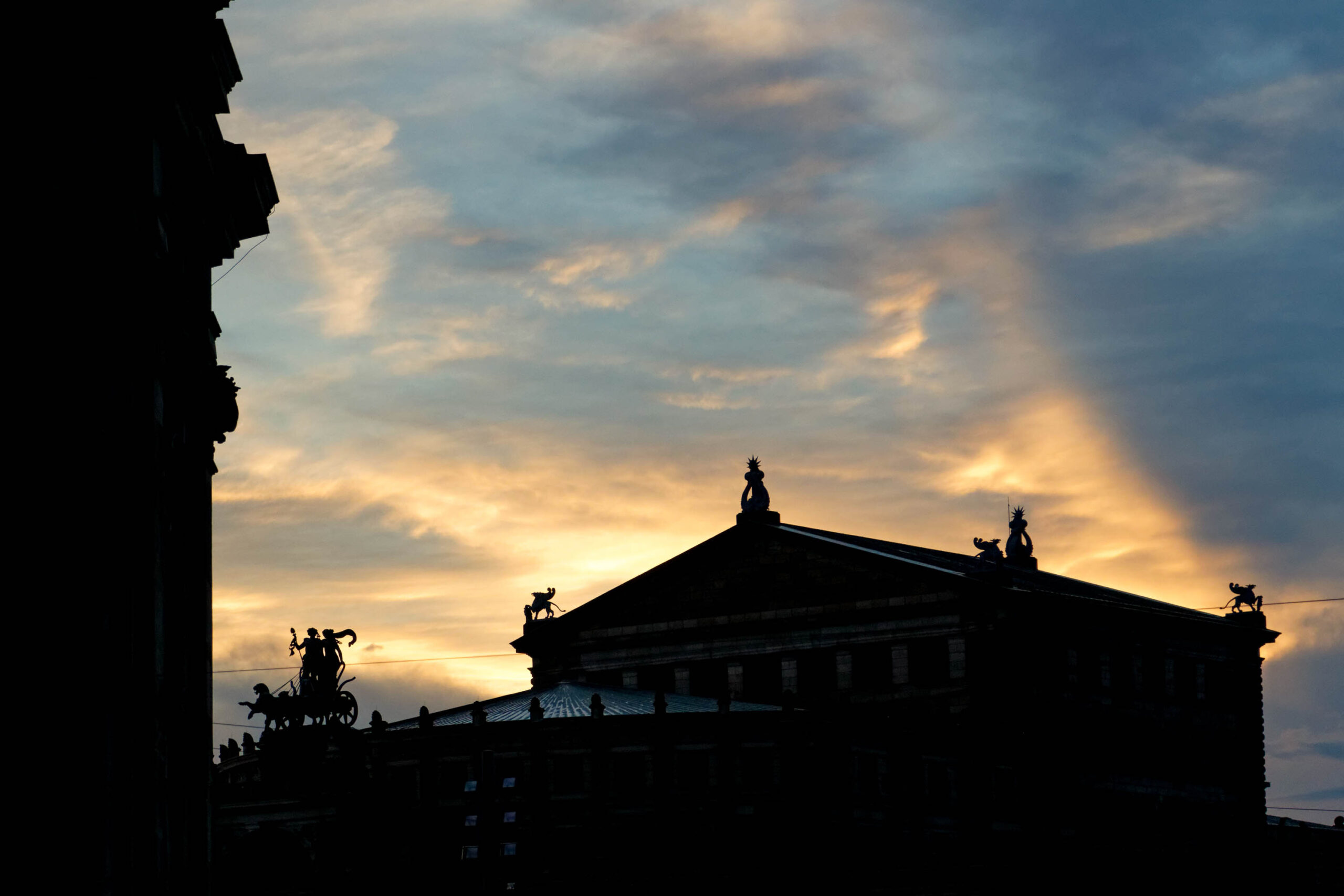 Das Bild zeigt eine Silhouettenaufnahme der Semperoper in Dresden, aufgenommen im August 2013. Der Himmel ist von einem dramatischen Sonnenuntergang in Gelb- und Orangetönen dominiert, der einen starken Kontrast zur dunklen Silhouette des Gebäudes bildet. Die filigranen Details der Architektur der Semperoper, einschließlich der Skulpturen auf dem Dach und der Säulen, sind deutlich erkennbar. Im Vordergrund ist ein Teil eines weiteren Gebäudes mit Skulpturen zu sehen, das die Komposition ergänzt. Die Aufnahme fängt die majestätische Erscheinung der Semperoper in einem stimmungsvollen Licht ein.