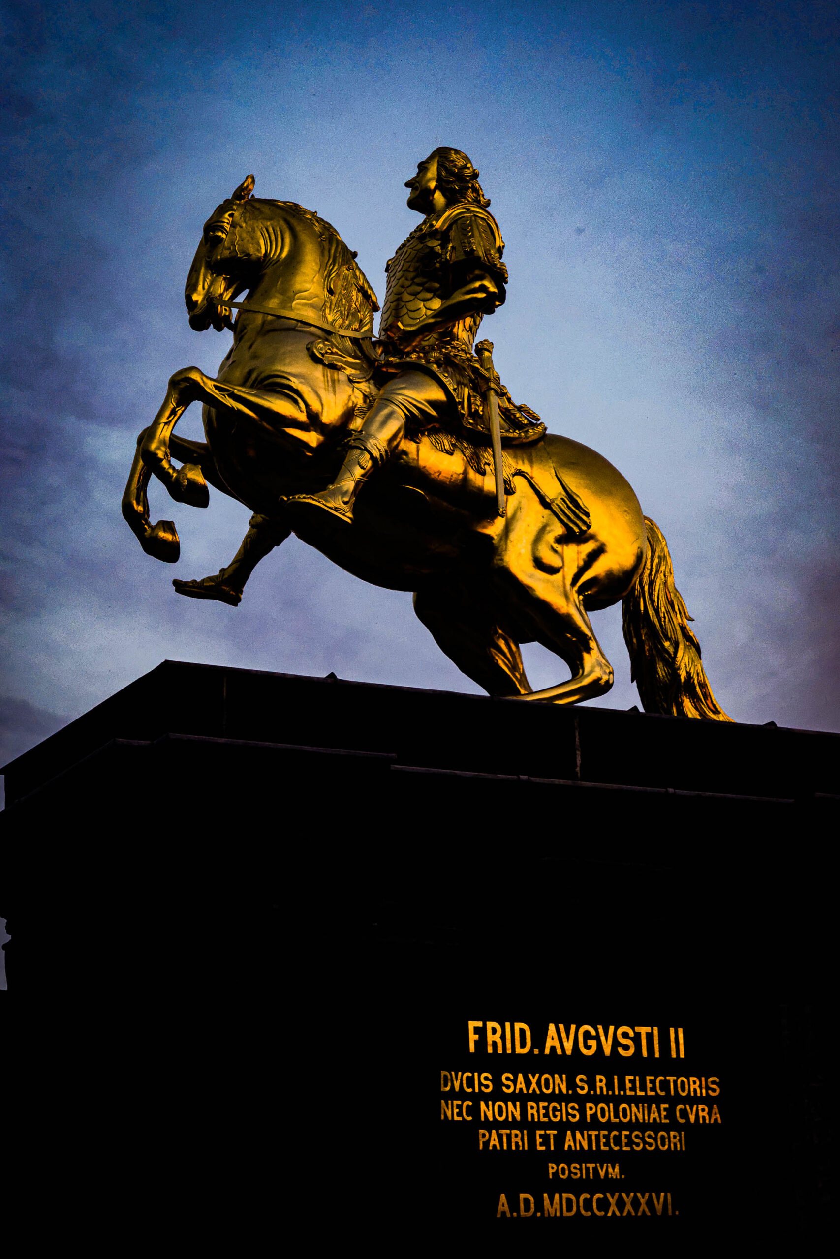 Das Bild zeigt eine detaillierte Nahaufnahme der Reiterstatue König Augusts des Starken, die sich auf dem Augustusbrunnen in Dresden befindet. Die Statue ist in Gold gehalten und steht vor einem dunklen Nachthimmel. Die Beleuchtung der Statue erzeugt einen starken Kontrast zum dunklen Hintergrund. Die Inschrift am Sockel der Statue ist lesbar. Die Szene vermittelt eine Atmosphäre von Größe, Macht und historischer Bedeutung.
