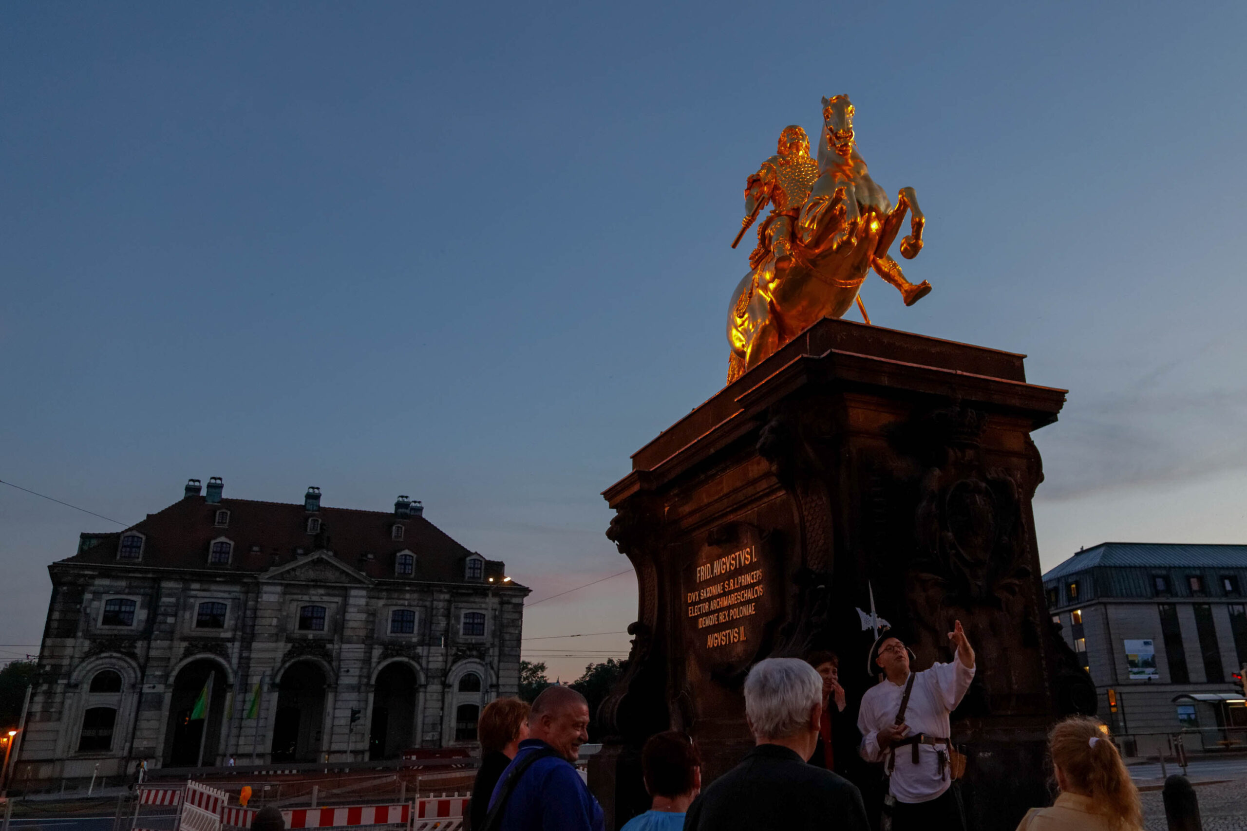 Die Aufnahme zeigt den Reiterstandbild des Augusts des Starken vor dem Zwinger in Dresden. Das Standbild ist golden und steht auf einem hohen Sockel. Im Hintergrund ist der Zwinger mit seiner beeindruckenden Architektur zu sehen. Mehrere Personen befinden sich vor dem Standbild und beobachten es. Die Szene ist von einer dunklen Nacht beleuchtet, wobei das Standbild und der Zwinger durch Scheinwerfer in Szene gesetzt werden. Baugerüste sind im Vordergrund sichtbar, was auf laufende Restaurierungsarbeiten hindeutet. Die Stimmung ist feierlich und respektvoll, aber auch von einer gewissen Melancholie geprägt, da die Aufnahme im August 2013 aufgenommen wurde, kurz nach der Flutkatastrophe.
