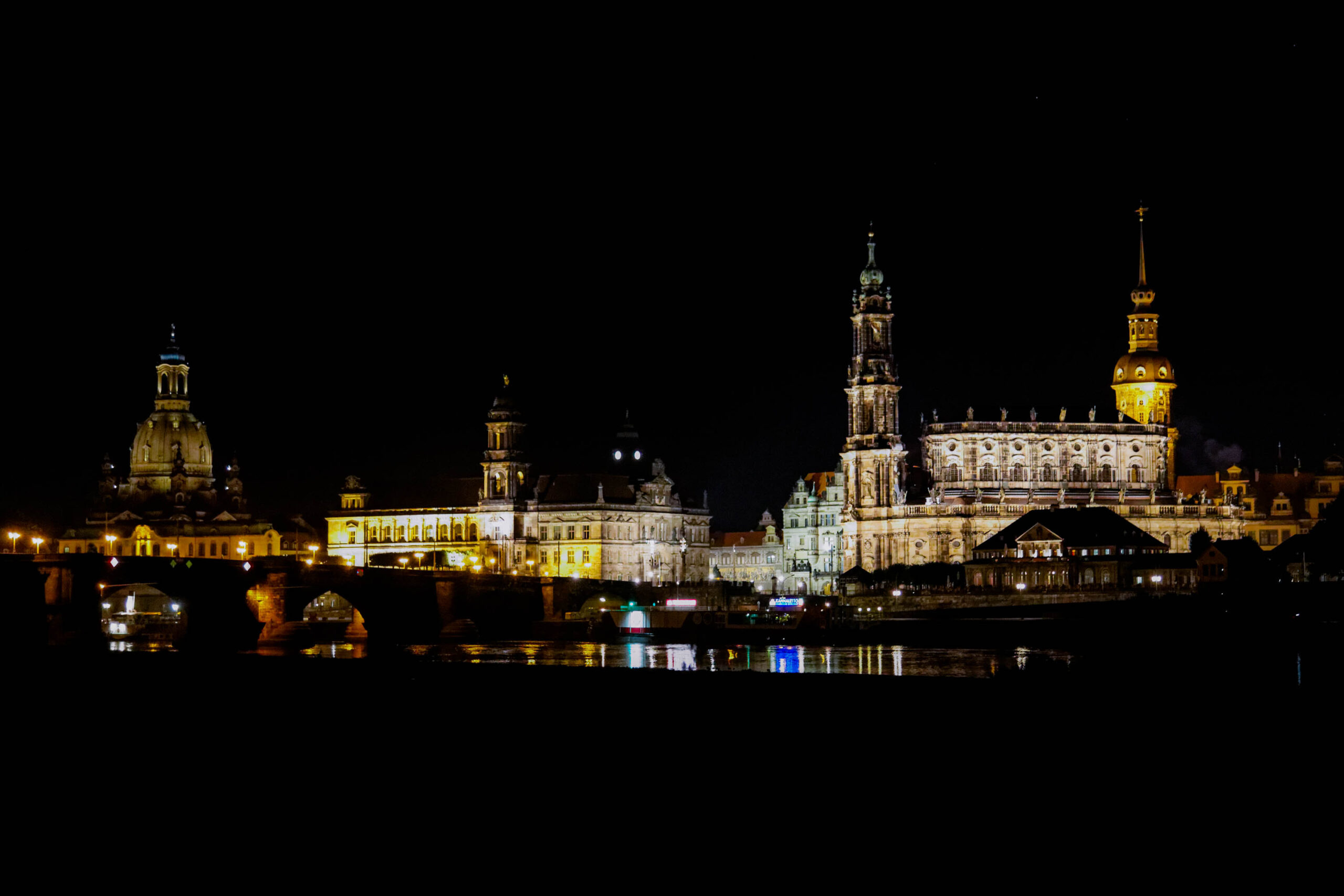 Die Aufnahme zeigt eine atemberaubende Nachtaufnahme der Dresdner Frauenkirche und des Residenzschlosses, aufgenommen vom Elbufer. Die Gebäude sind wunderschön beleuchtet, wobei die goldenen und weißen Lichter die architektonischen Details hervorheben. Die Elbe spiegelt die Lichter der Gebäude wider und erzeugt einen magischen Effekt. Der Himmel ist dunkel und klar, was die Beleuchtung der Gebäude noch verstärkt. Die Aufnahme fängt die Schönheit und den Charme Dresdens bei Nacht ein.