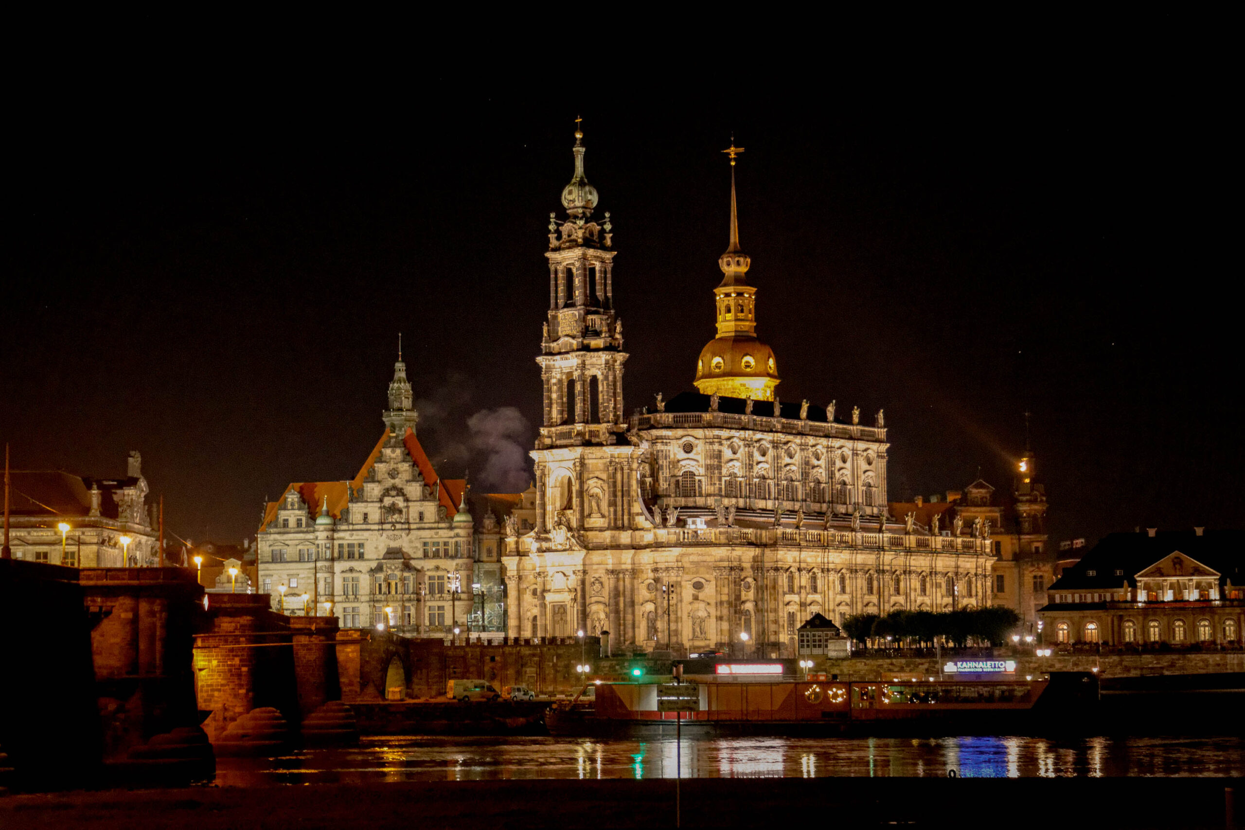 Die Aufnahme zeigt die Frauenkirche in Dresden bei Nacht. Die Kirche ist von warmen, goldenen Lichtern beleuchtet, die ihre Fassade hervorheben und einen starken Kontrast zum dunklen Nachthimmel bilden. Die Beleuchtung betont die architektonischen Details der Kirche, einschließlich der Kuppel und der Türme. Die Elbe, die vor der Kirche fließt, spiegelt das Licht der Kirche und der umliegenden Gebäude wider, was eine malerische und fast surreale Atmosphäre schafft. Im Vordergrund ist ein Teil einer Brücke sichtbar, die das Ufer verbindet. Die umliegenden Gebäude sind ebenfalls beleuchtet, aber in einem gedämpfteren Ton, was die Frauenkirche als das Hauptaugenmerk der Szene hervorhebt. Die Aufnahme vermittelt ein Gefühl von Erhabenheit, Schönheit und historischer Bedeutung.