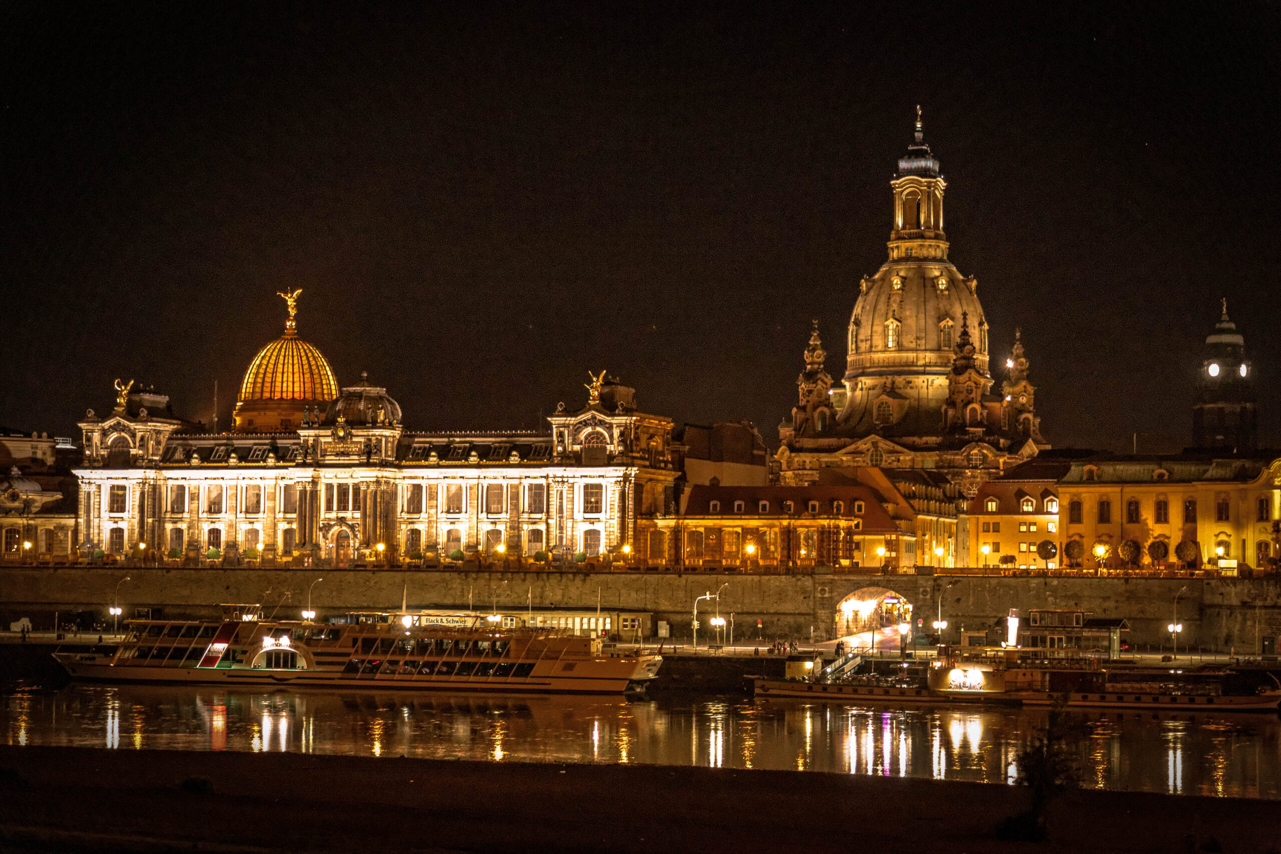 Die Aufnahme zeigt eine atemberaubende Nachtansicht des Dresdner Zwingers und der Frauenkirche. Der Zwinger, mit seiner beeindruckenden Fassade und dem goldenen Kuppel, wird durch zahlreiche Lichter in Szene gesetzt. Die Frauenkirche, ebenfalls beleuchtet, dominiert den Hintergrund. Die Spiegelung der Gebäude im Flusswasser verstärkt die magische Atmosphäre. Mehrere Schiffe liegen am Ufer vor dem Zwinger. Der Himmel ist dunkel und klar, was die Beleuchtung der Gebäude hervorhebt.