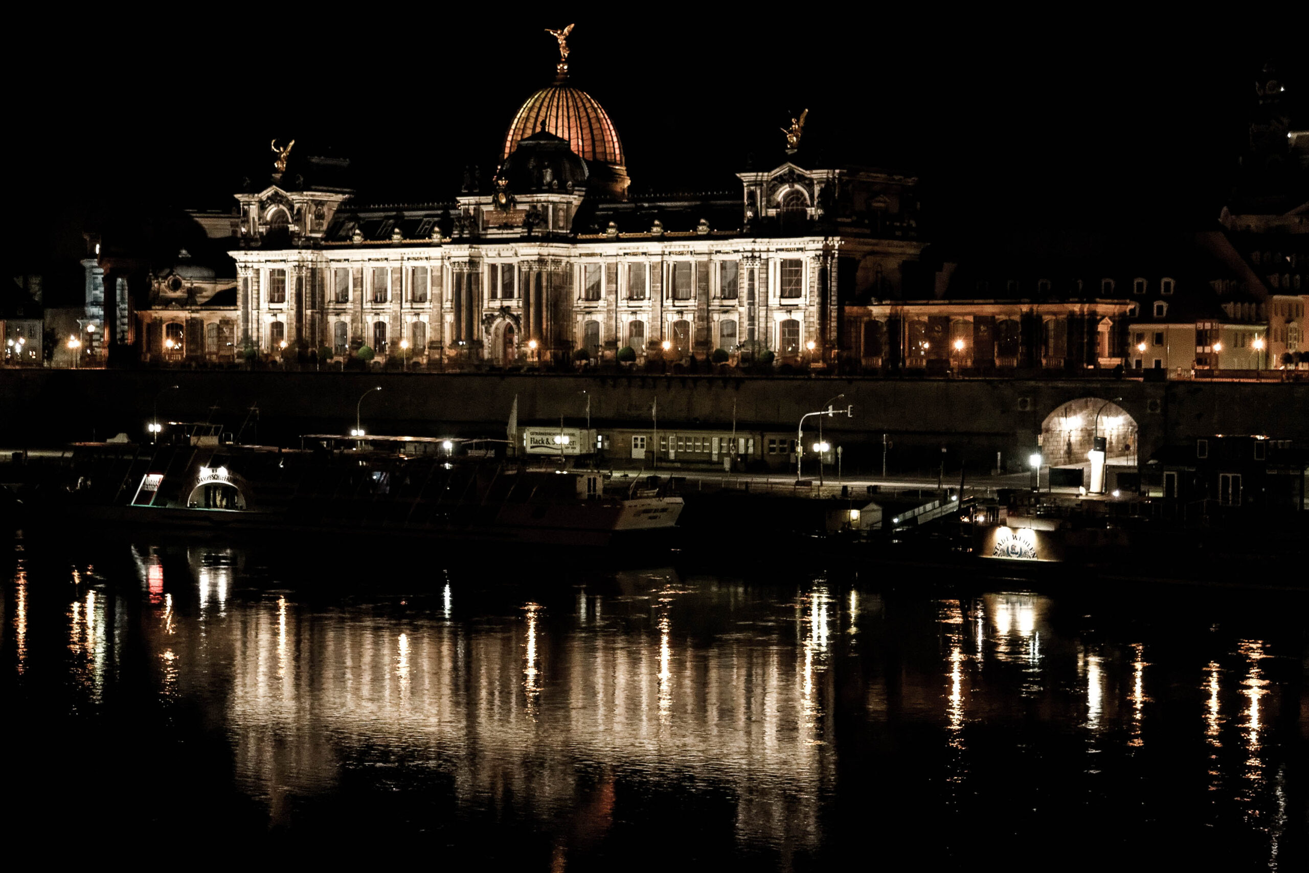 Die Aufnahme zeigt den Zwinger in Dresden bei Nacht. Das Wasser der Elbe spiegelt die Beleuchtung des Palastes wider, was eine stimmungsvolle Atmosphäre erzeugt. Die Aufnahme ist in einem dunklen Farbton gehalten, was die Beleuchtung des Palastes hervorhebt. Im Vordergrund sind einige Schiffe und Uferstrukturen zu sehen.