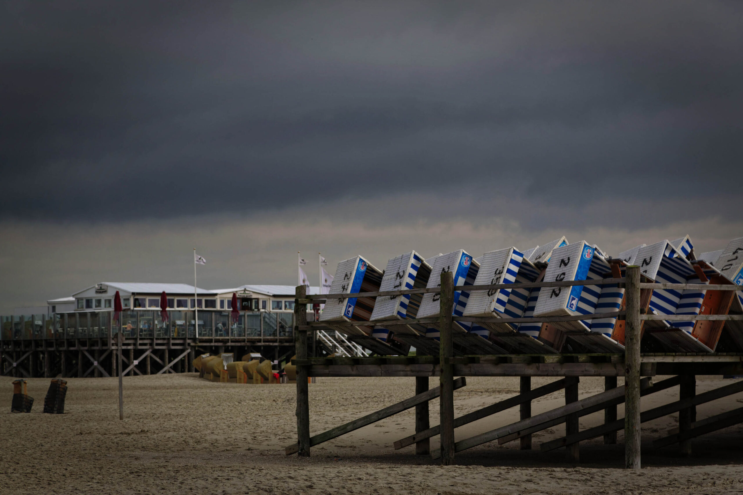 Das Foto fängt eine Szene am Strand von St. Peter-Ording ein, einem Küstenort an der Nordsee. Im Vordergrund stehen mehrere Strandkörbe, die in verschiedenen Farben und Mustern gestreift sind. Sie sind auf einer Holzkonstruktion platziert, die vermutlich Teil einer Promenadenanlage ist. Im Hintergrund sind Strandhäuser und eine Promenade zu sehen, die auf Stelzen errichtet sind. Der Himmel ist dunkel und bewölkt, was eine melancholische Stimmung erzeugt. Einige Personen sind im Hintergrund zu sehen, die sich auf der Promenade bewegen.