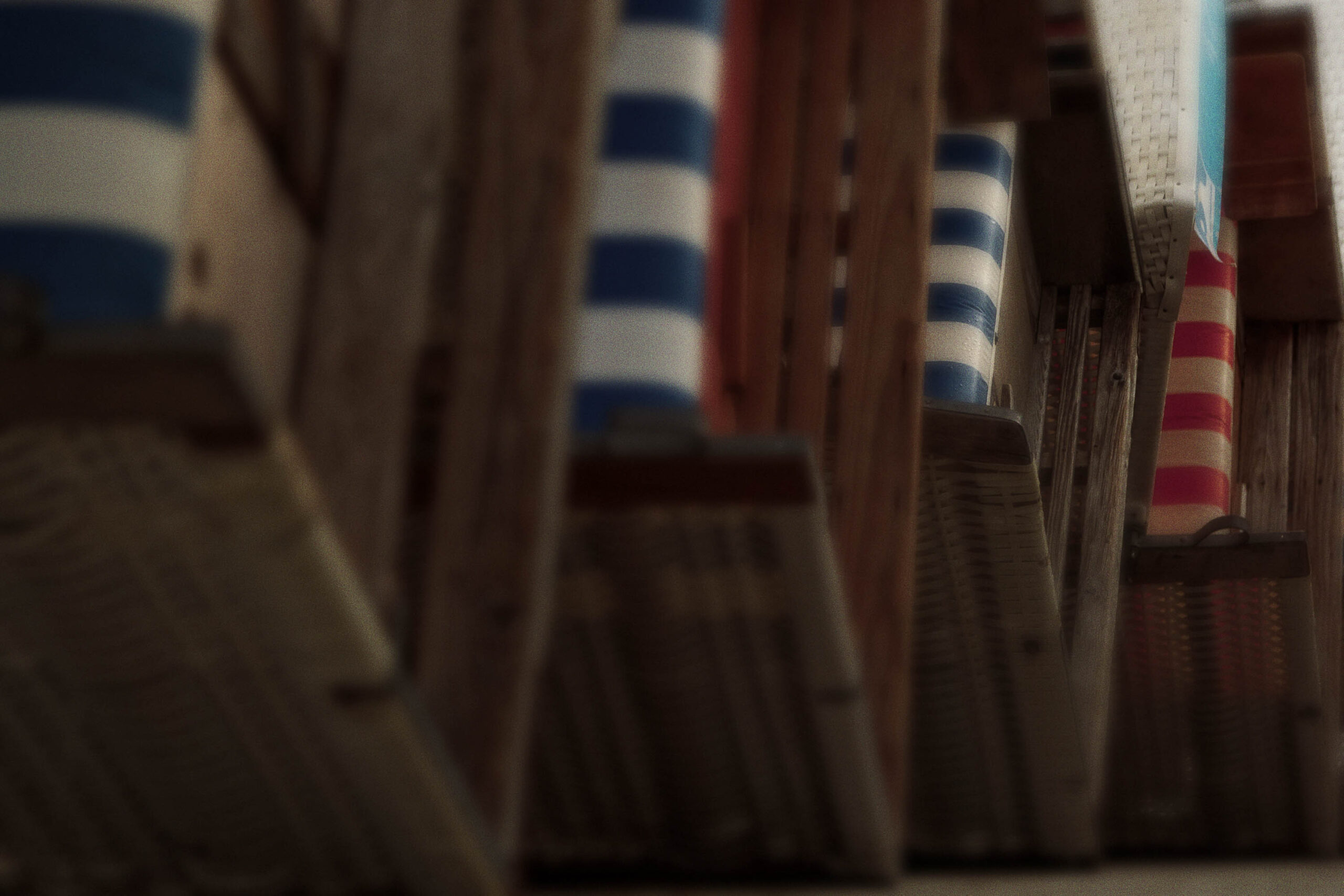 Das Bild zeigt eine ungewöhnliche Perspektive auf eine Reihe von Holzpfählen, die in einem Muster aus roten und weißen Streifen bemalt sind. Die Pfähle scheinen sich in der Ferne zu erheben und bilden eine geometrische Struktur. Der Hintergrund ist verschwommen und deutet auf einen Strand und einen Himmel hin. Die Bildkomposition ist interessant, da sie eine ungewöhnliche Perspektive und eine ungewöhnliche Komposition verwendet. Das Bild ist in einem dunklen Ton gehalten, was ihm eine düstere und geheimnisvolle Atmosphäre verleiht.