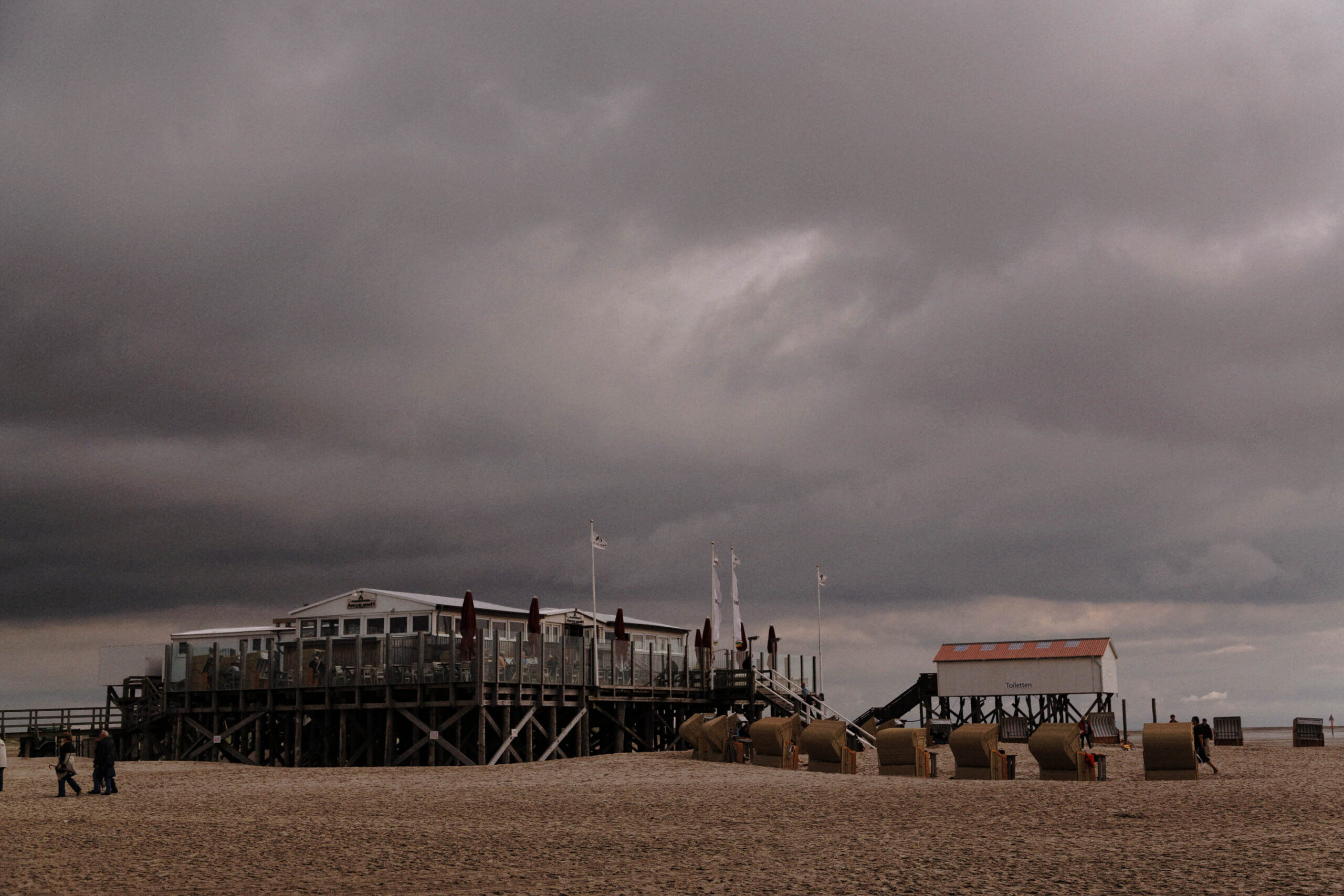 Die Aufnahme zeigt eine Szene am Strand von St. Peter-Ording an der Nordsee. Im Vordergrund erstreckt sich ein breiter Sandstrand, der von einem dunklen, bedrohlichen Himmel bedeckt ist. Ein markantes Merkmal ist ein erhöhter Strandkorb-Pavillon auf Stelzen, der mehrere Etagen und eine Terrasse umfasst. Im Hintergrund befindet sich ein kleines, rotes Gebäude mit einem Satteldach. Mehrere gestapelte Strandkörbe sind ebenfalls sichtbar. Einige Personen sind im Bild zu erkennen, die sich auf dem Strand aufhalten. Die Szene vermittelt eine Atmosphäre von Ruhe und Melancholie, verstärkt durch das düstere Wetter.