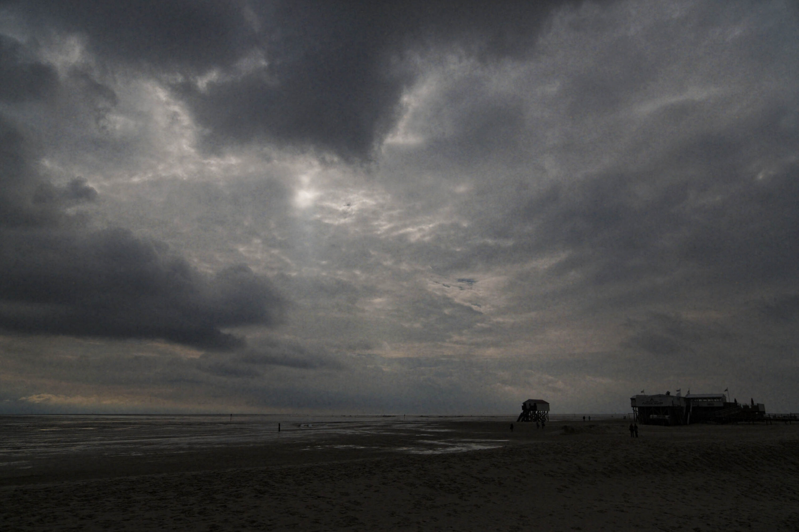 Die Aufnahme zeigt einen weiten Strandabschnitt, der von dunklem Sand bedeckt ist. Im Vordergrund erstreckt sich eine flache Wasserfläche, die sich bis zum Horizont erstreckt. Am Horizont sind Gebäude und Strukturen erkennbar, die wahrscheinlich zu St. Peter-Ording gehören. Der Himmel ist von dunklen, bedrohlich wirkenden Wolken bedeckt, die eine düstere und stürmische Atmosphäre erzeugen. Einige Personen sind auf dem Strand zu sehen, die die Weite und die Atmosphäre des Ortes unterstreichen.