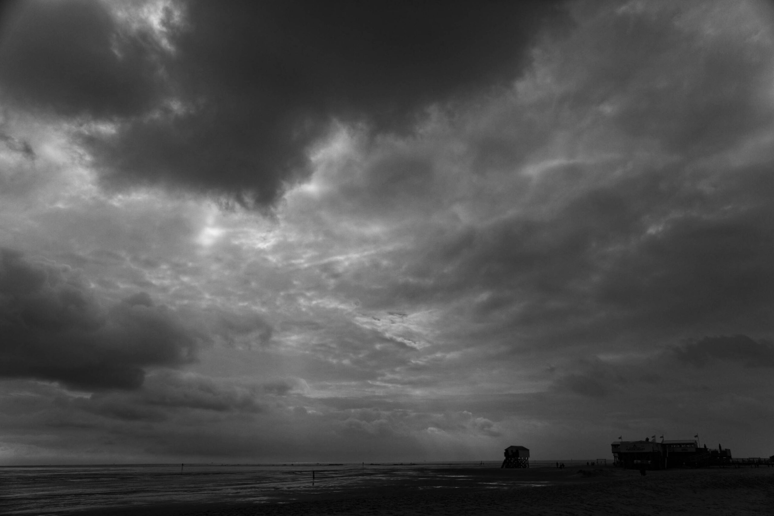 Das Schwarz-Weiß-Foto fängt die raue Schönheit von St. Peter-Ording an der Nordsee ein. Ein dramatischer Himmel dominiert das Bild, mit dunklen Wolken, die sich über das Meer erstrecken. Im Vordergrund liegt ein breiter Sandstrand, der von den Wellen des Meeres umspült wird. Im Hintergrund sind zwei markante Strukturen zu erkennen: ein hölzerner Steg und ein hölzerner Turm, die typisch für die Küstenarchitektur der Region sind. Das Licht ist gedämpft und erzeugt eine melancholische Stimmung.