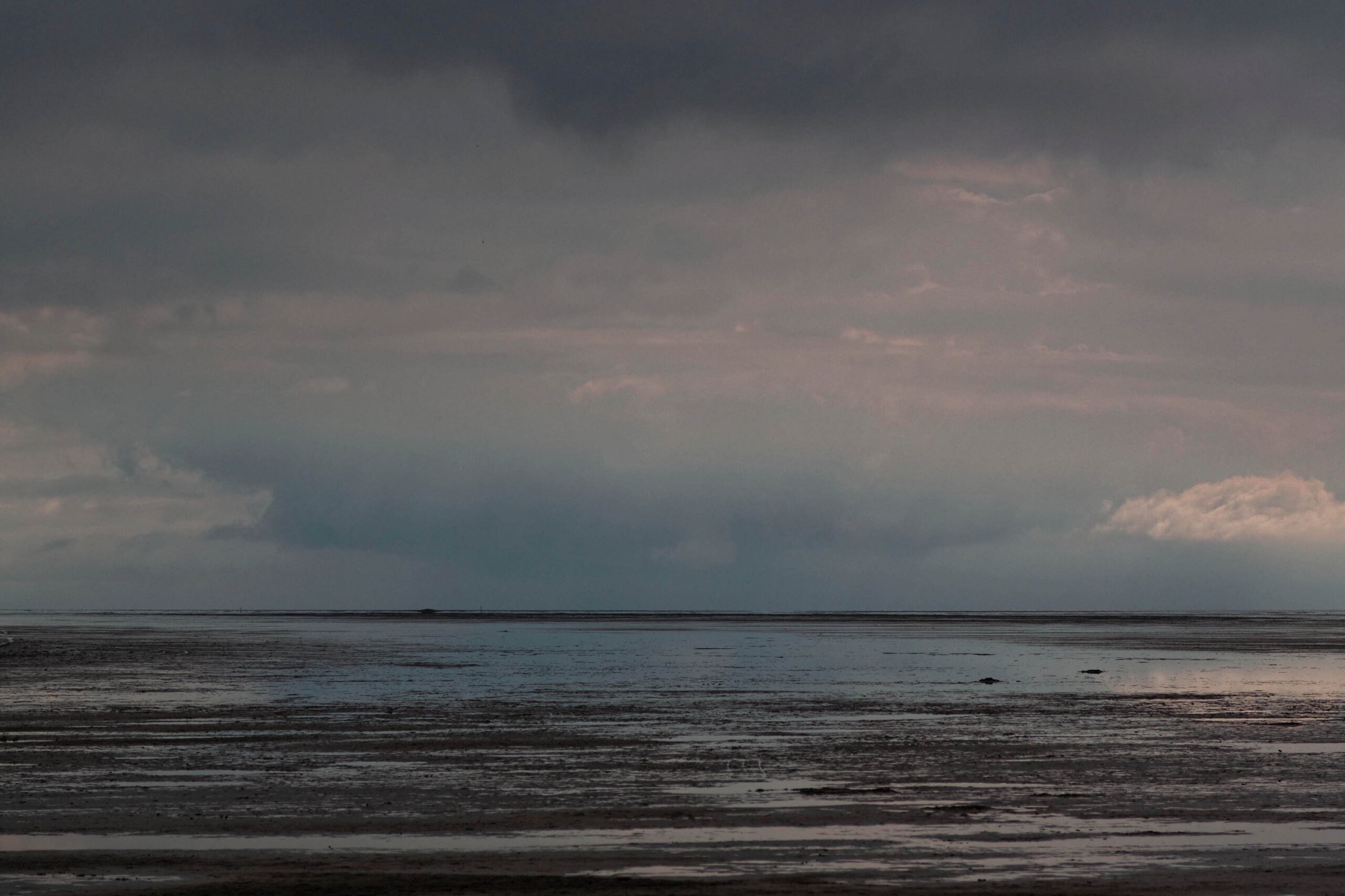 Die Aufnahme zeigt eine typische Landschaft der Nordsee, vermutlich in der Nähe von St. Peter-Ording. Der Fokus liegt auf der Weite des Strandes und des Himmels. Die Farben sind gedämpft und erzeugen eine ruhige, fast melancholische Stimmung. Die Reflexionen im nassen Sand verstärken den Eindruck von Weite und Unendlichkeit.