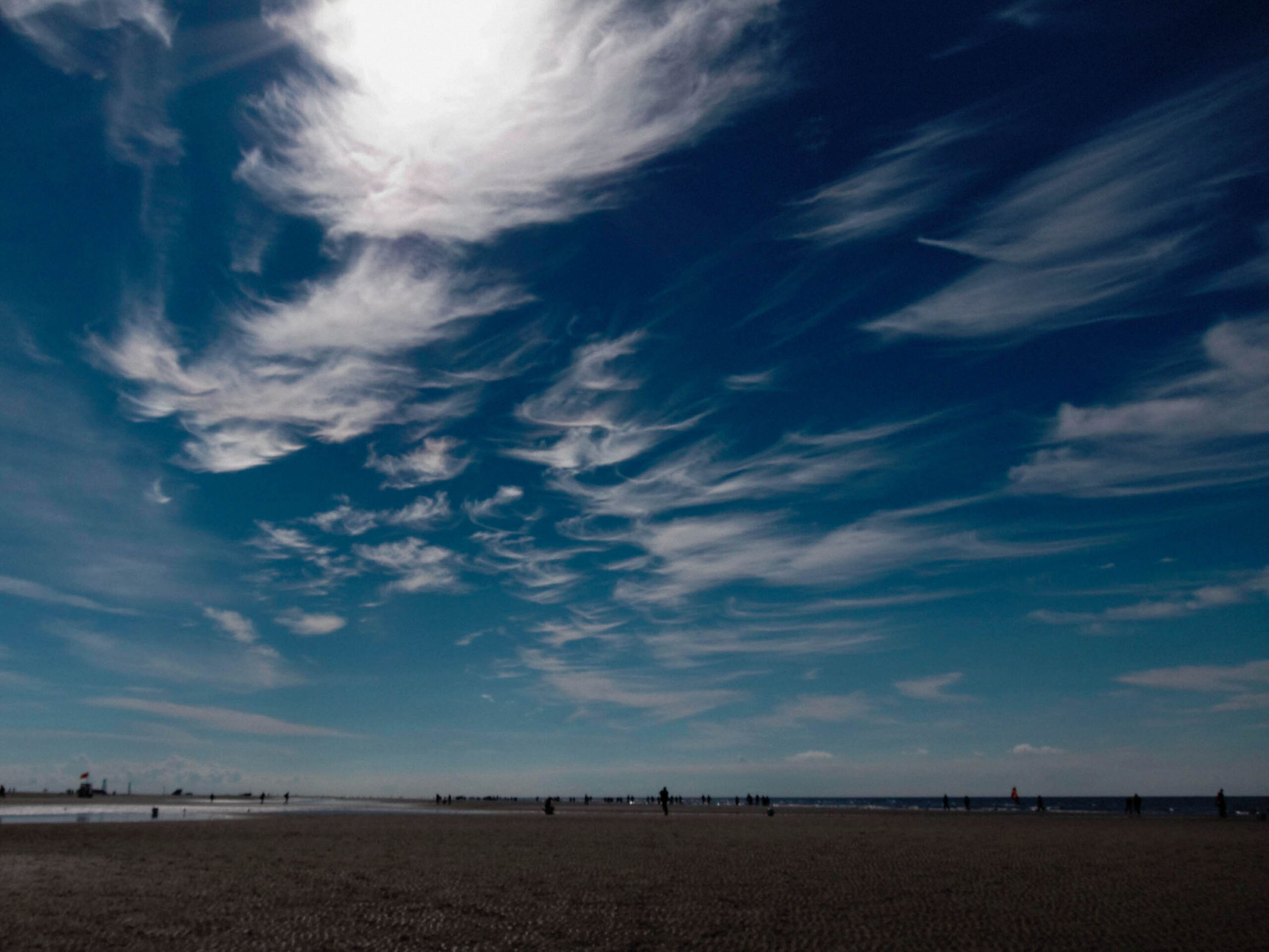 Die Aufnahme zeigt einen weiten Blick auf einen Sandstrand, vermutlich in St. Peter-Ording an der Nordsee. Der Himmel ist tiefblau und von zarten, faserartigen Wolken bedeckt, die sich in Richtung des Horizonts ziehen. Der Sandstrand ist dunkel und fast vollständig von Silhouetten von Menschen und einigen wenigen Objekten besetzt. Das Meer ist in der Ferne sichtbar, ebenfalls als dunkle Silhouette dargestellt. Die Szene vermittelt eine ruhige und friedliche Atmosphäre.