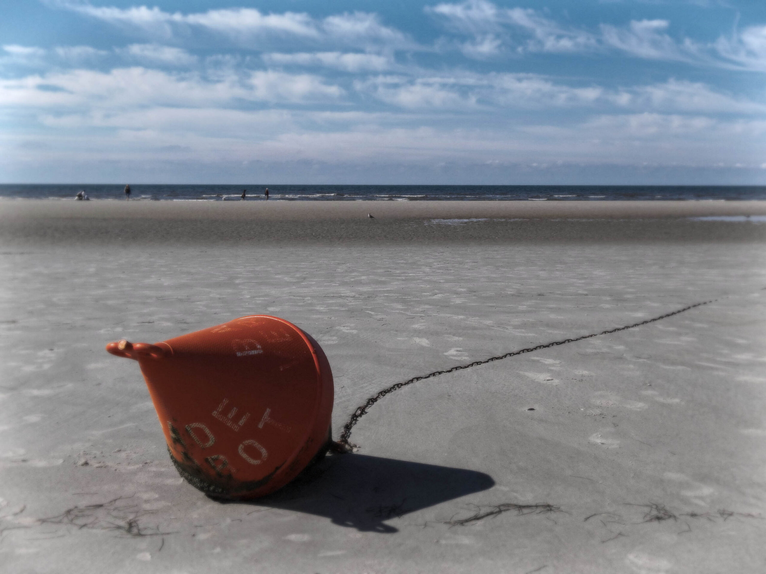 Das Foto zeigt eine orangefarbene Boje, die im feuchten Sand eines Strandes liegt. Die Boje ist rund und hat eine deutliche Kette, die ins Meer führt. Im Hintergrund erstreckt sich ein weiter Sandstrand, auf dem sich einige Personen aufhalten, die im Wasser spielen oder spazieren gehen. Der Himmel ist bewölkt, was eine diffuse Beleuchtung erzeugt. Die Szene vermittelt eine ruhige und friedliche Atmosphäre.