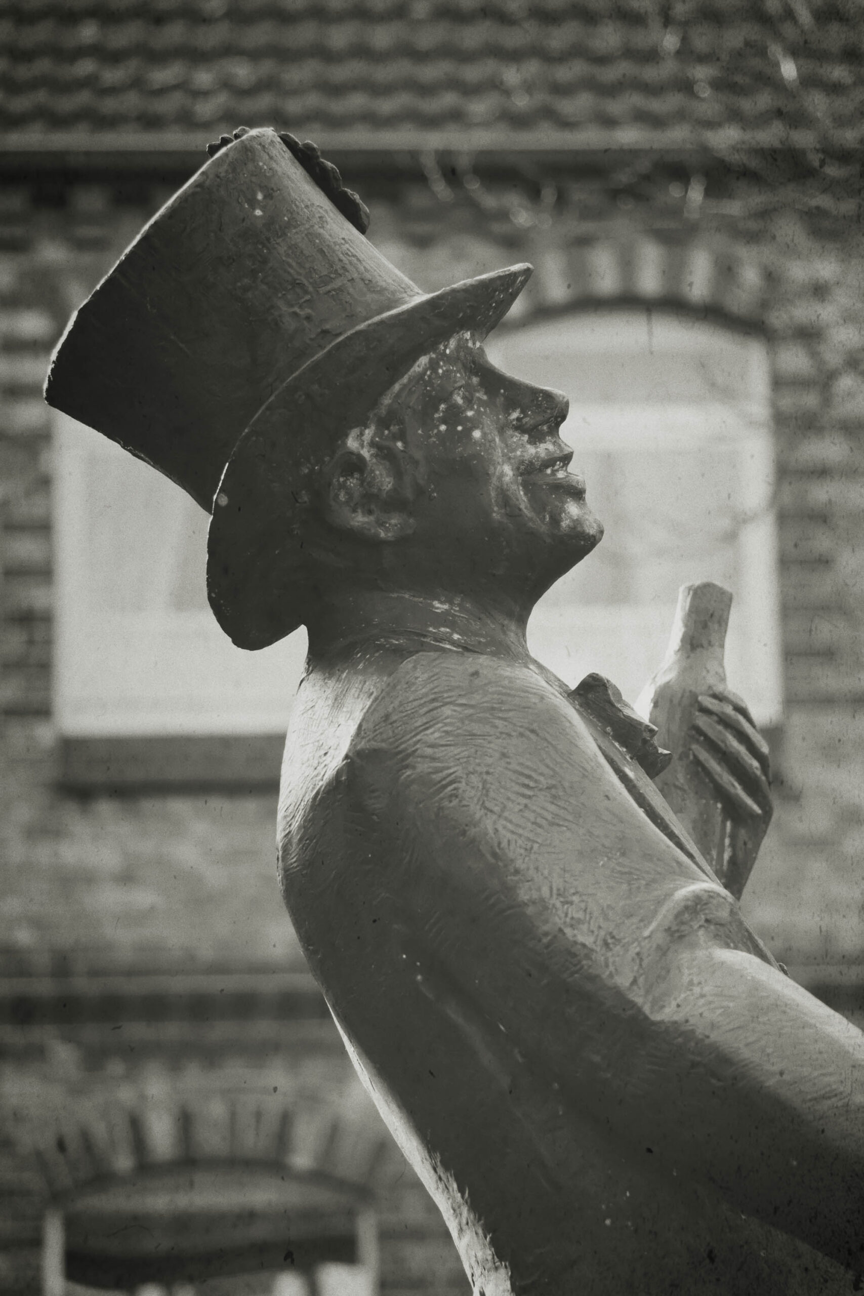 Die Fotografie zeigt eine detaillierte Statue eines Mannes, vermutlich aus Bronze oder einem ähnlichen Material. Er trägt einen hochgezogenen Zylinderhut und eine Art Jackett oder Anzug. In seiner rechten Hand hält er eine Flasche, die er möglicherweise trinkt oder anbietet. Die Statue ist in einem dynamischen Profil dargestellt, das einen Eindruck von Bewegung und Persönlichkeit vermittelt. Der Hintergrund besteht aus einem fragmentarischen Blick auf ein Gebäude, das die Aufmerksamkeit auf die Statue lenkt. Die Schwarz-Weiß-Darstellung verstärkt die Textur und die Details der Statue und verleiht der Szene eine zeitlose Qualität.