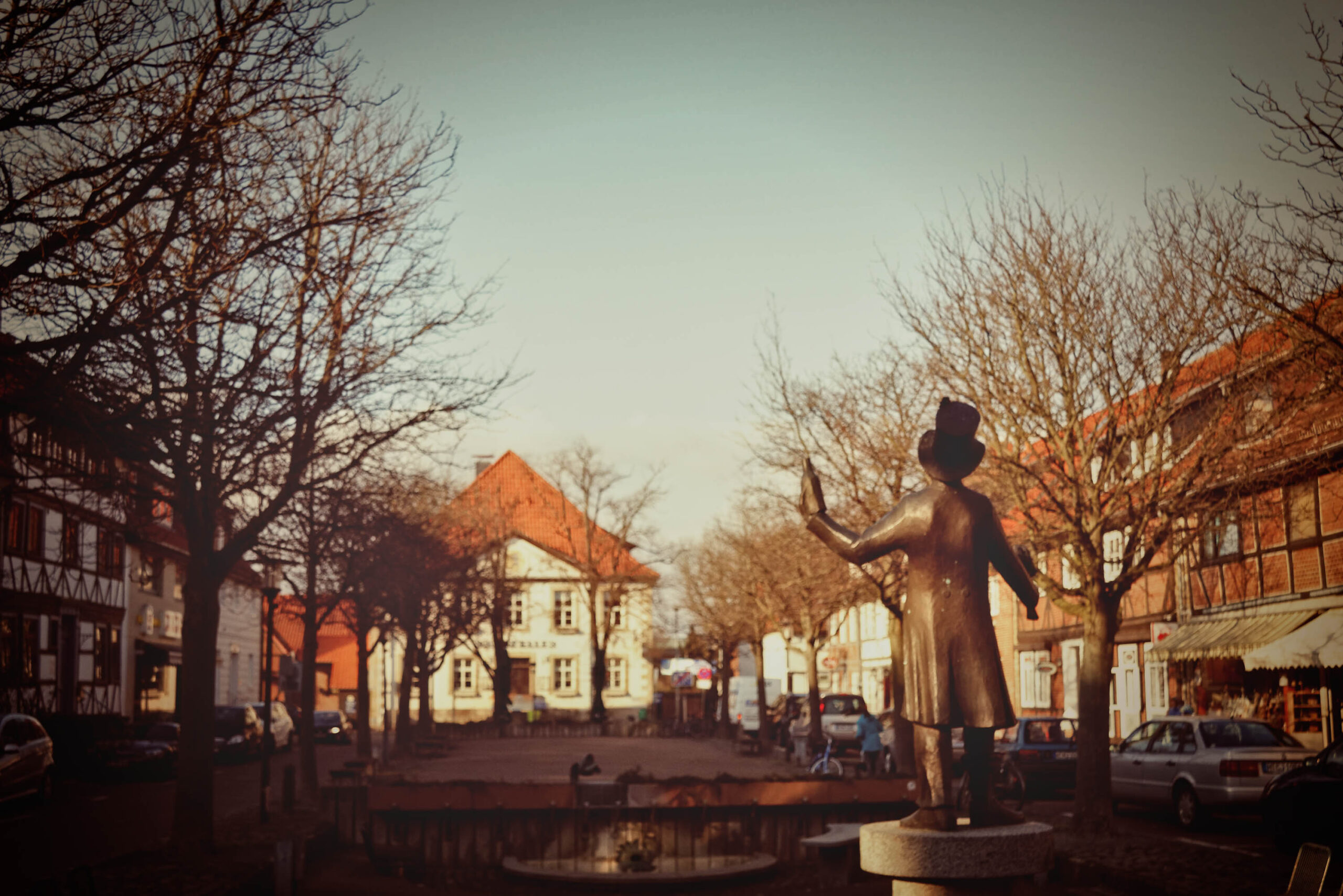 Das Foto zeigt eine Bronzestatue einer Frau, die auf einem Sockel vor einem historischen Stadtbild steht. Die Frau hat ihre Arme erhoben, als ob sie etwas präsentiert oder begrüßt. Im Hintergrund sind Fachwerkhäuser mit roten Dächern zu sehen, die typisch für die Region sind. Der Himmel ist hell und scheint auf die Szene zu fallen, was eine warme und nostalgische Atmosphäre erzeugt. Die Szene wirkt ruhig und friedlich, als ob die Zeit stehen geblieben wäre.