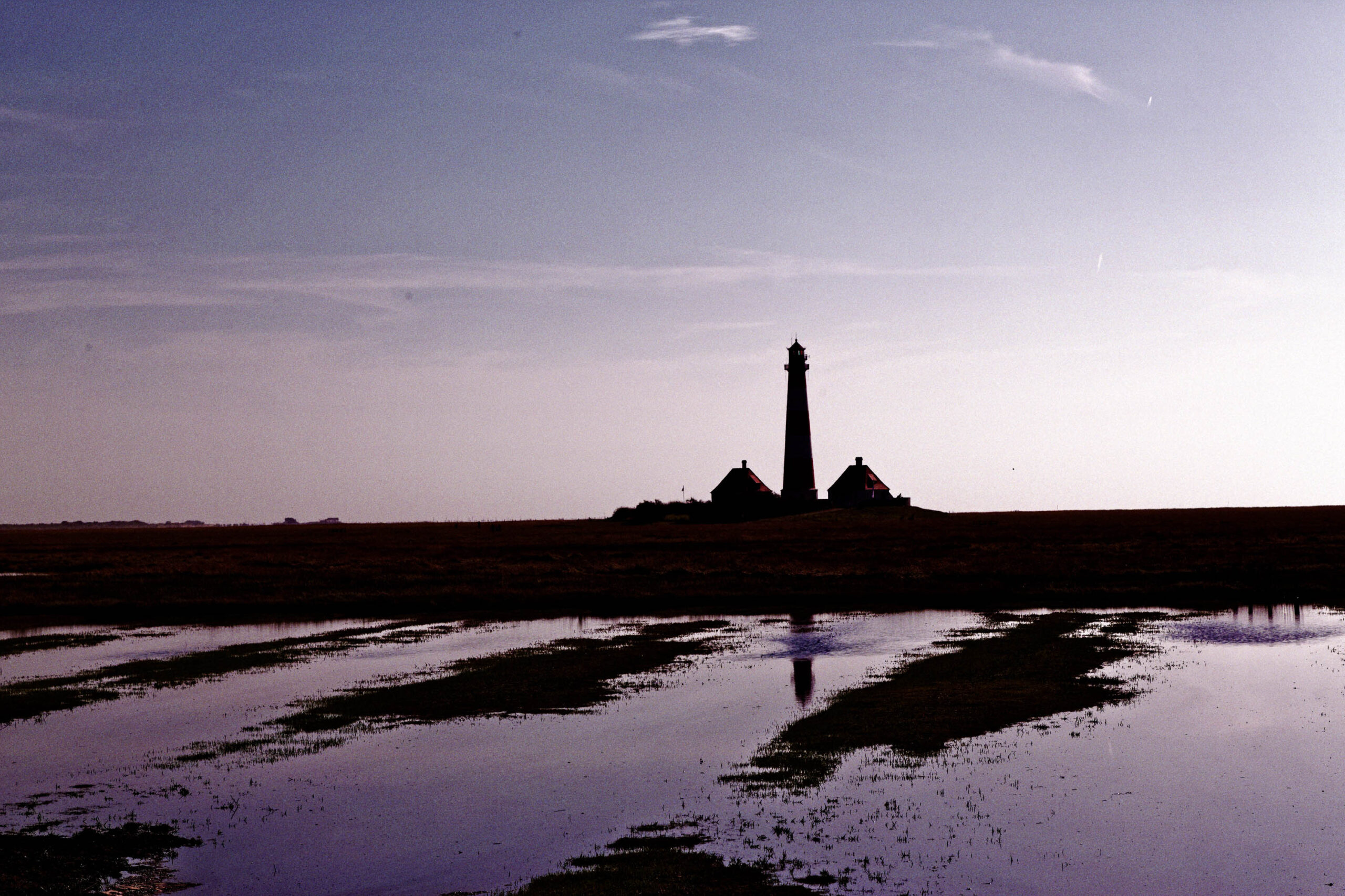 Die Aufnahme zeigt eine dramatische Silhouettenaufnahme des Westerhever Leuchtturms, der sich vor einem violetten Himmel erhebt. Die Landschaft im Vordergrund besteht aus einer flachen, feuchten Ebene, die sich im Wasser spiegelt. Die Szene vermittelt eine ruhige, fast mystische Atmosphäre. Der Leuchtturm und die umliegenden Gebäude sind scharf abgegrenzt und bilden einen starken Kontrast zum Himmel. Die Reflexionen im Wasser verstärken die Tiefe und den surrealen Charakter des Bildes.
