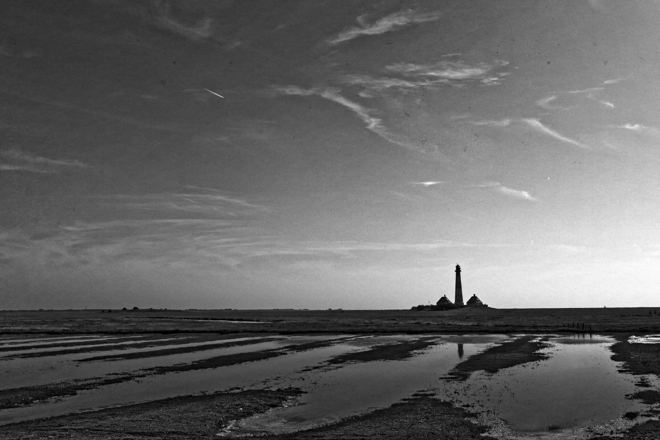 Das Schwarz-Weiß-Foto zeigt eine weite Landschaft, die von einer Wasserfläche dominiert wird, vermutlich eine Gezeitengebiet oder eine flache Bucht. Im Hintergrund erhebt sich der Westerhever Leuchtturm, dessen Silhouette deutlich erkennbar ist. Die Landschaft ist von einem Himmel mit deutlichen Wolkenformationen überzogen. Die Wasserfläche reflektiert das Licht und die Wolken, was eine ruhige und fast surreale Atmosphäre erzeugt. Die Textur des Bodens ist durch die Wasserlinien und die Reflexionen betont. Die Komposition ist einfach, aber effektiv, mit dem Leuchtturm als zentrales Element, das den Blick des Betrachters lenkt.