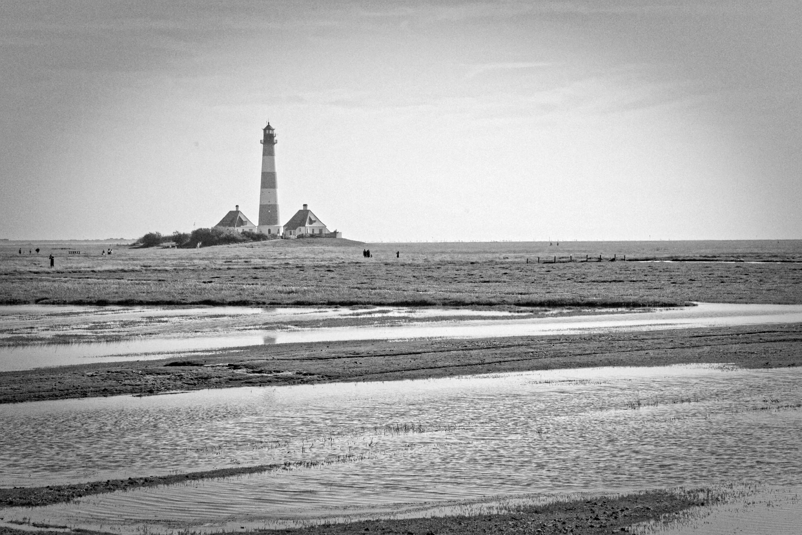 Das Foto zeigt den Westerhever Leuchtturm, ein markantes Wahrzeichen an der Küste der Nordsee. Der Leuchtturm steht auf einer kleinen, künstlich angelegten Insel, die von flachem Wasser umgeben ist. Das Watt erstreckt sich bis zum Horizont. Einige Personen sind in einiger Entfernung zu sehen. Das Licht ist diffus, was eine ruhige und friedliche Atmosphäre erzeugt. Die Textur des Wassers und des Bodens ist deutlich erkennbar.